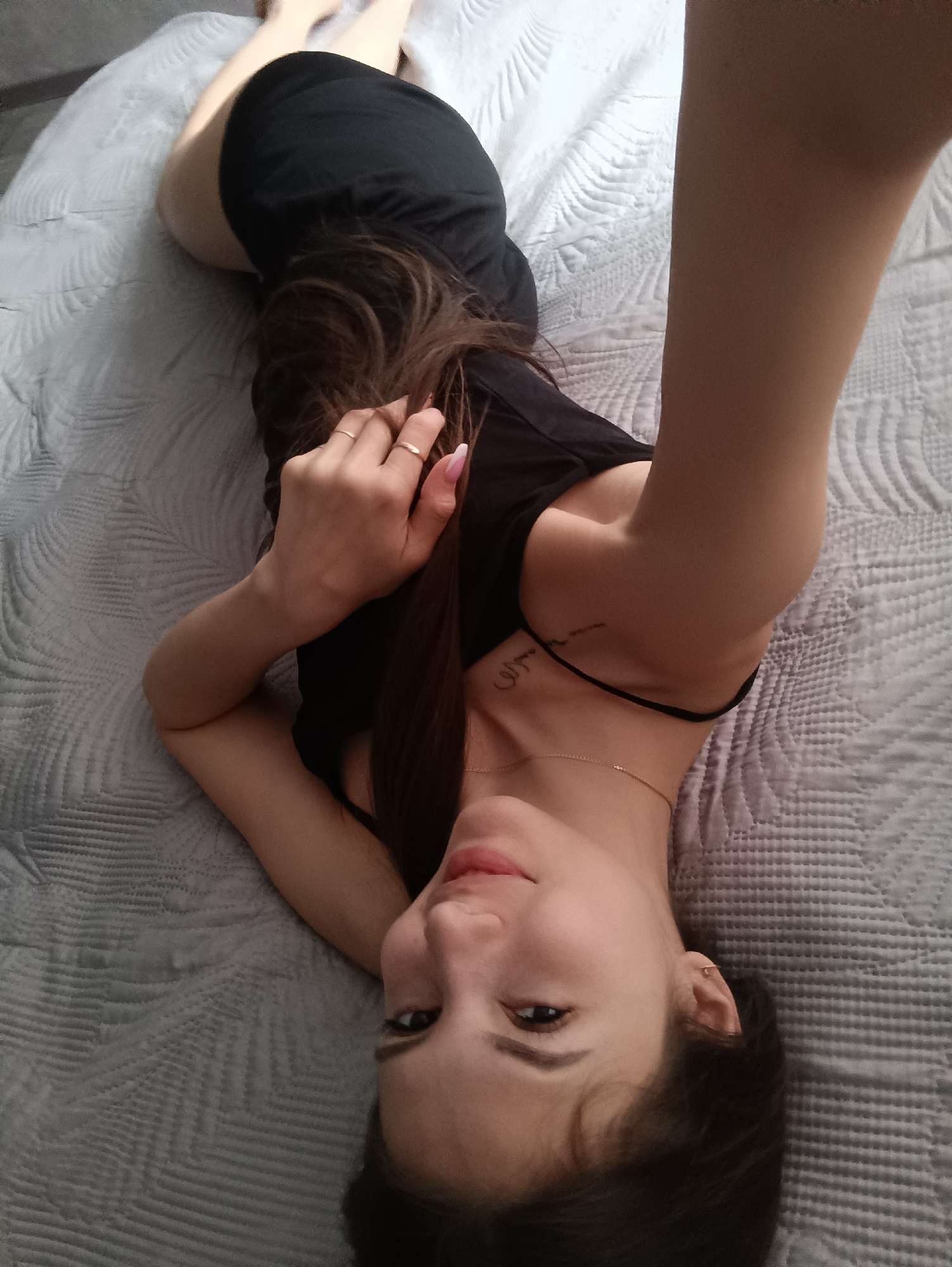 A young woman lies on a gray quilted bedspread, wearing a black dress. Her hair, dark brown, cascades onto the bed. She is holding her arm up, possibly taking a selfie. She has a tattoo on her upper chest/shoulder area and is wearing a delicate necklace. A small hoop earring is visible in her ear. She has rings on her fingers. The location appears to be indoors, likely a bedroom.