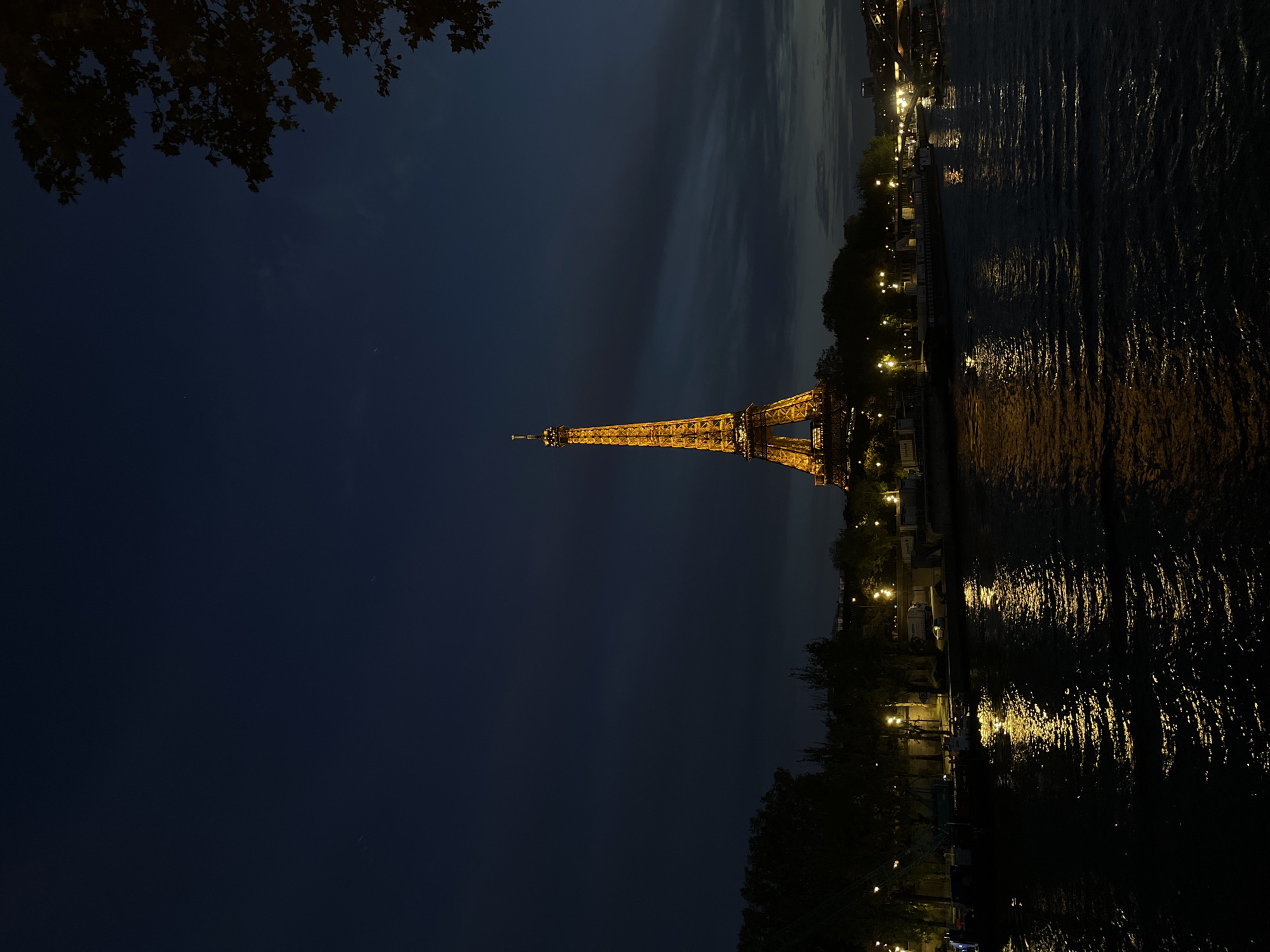 The image captures a nighttime view of the Eiffel Tower in Paris, France, illuminated in golden light. The iconic landmark stands prominently against a dark navy blue sky, which shows faint traces of clouds. In the foreground and to the right, a calm body of water, likely the Seine River, reflects the warm glows of numerous lights. These reflections create shimmering streaks of light on the water's surface, indicating movement or ripples.
Along the riverbank on the right, trees and what appear to be structures or buildings are dotted with bright streetlights, contributing to the reflections on the water. The upper left corner of the frame is obscured by dark foliage, possibly tree leaves, silhouetted against the night sky. The overall scene is serene and iconic, characteristic of an evening in Paris. The time of day is clearly night, with artificial lights providing the main illumination. There are no people distinctly visible, nor is any text present in the image.