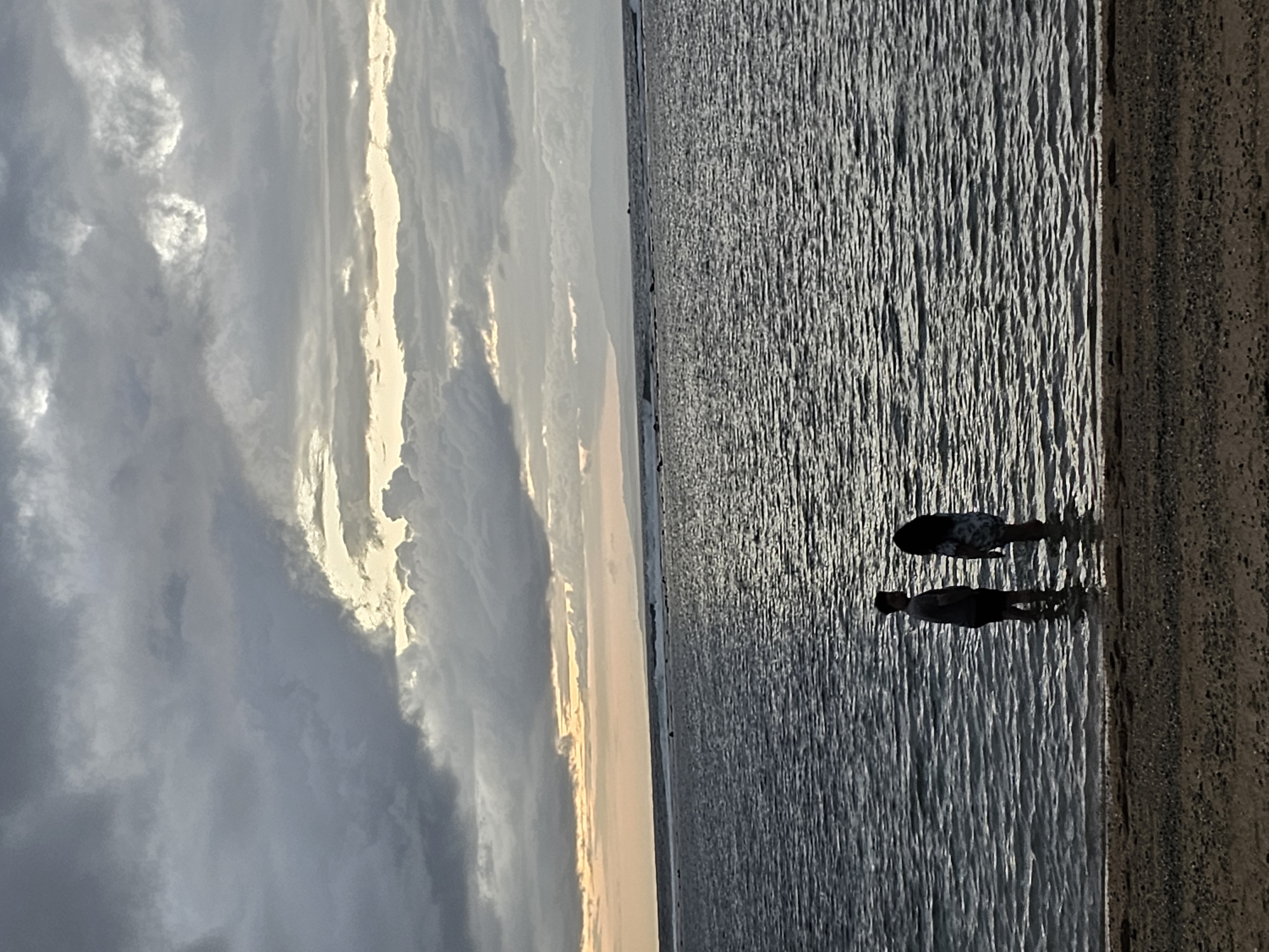 Two figures, one appearing male and the other female, stand partially submerged in shallow ocean water. The male figure is wearing a dark t-shirt and shorts, while the female figure is wearing a patterned dress or tunic. They are facing each other, suggesting an interaction, possibly a conversation. The water is calm with visible ripples. In the background, the ocean extends to the horizon where waves can be seen breaking further out. A distant landmass or island is visible on the horizon. The sky is overcast with dramatic, layered clouds, some of which are illuminated by sunlight breaking through. The foreground consists of a sandy beach with small pebbles. The overall lighting suggests it is either dawn or dusk. The scene is likely located along the coastline of Denpasar, Indonesia, given the presence of ocean, beach, and the tropical ambiance indicated by the vegetation on the distant landmass.