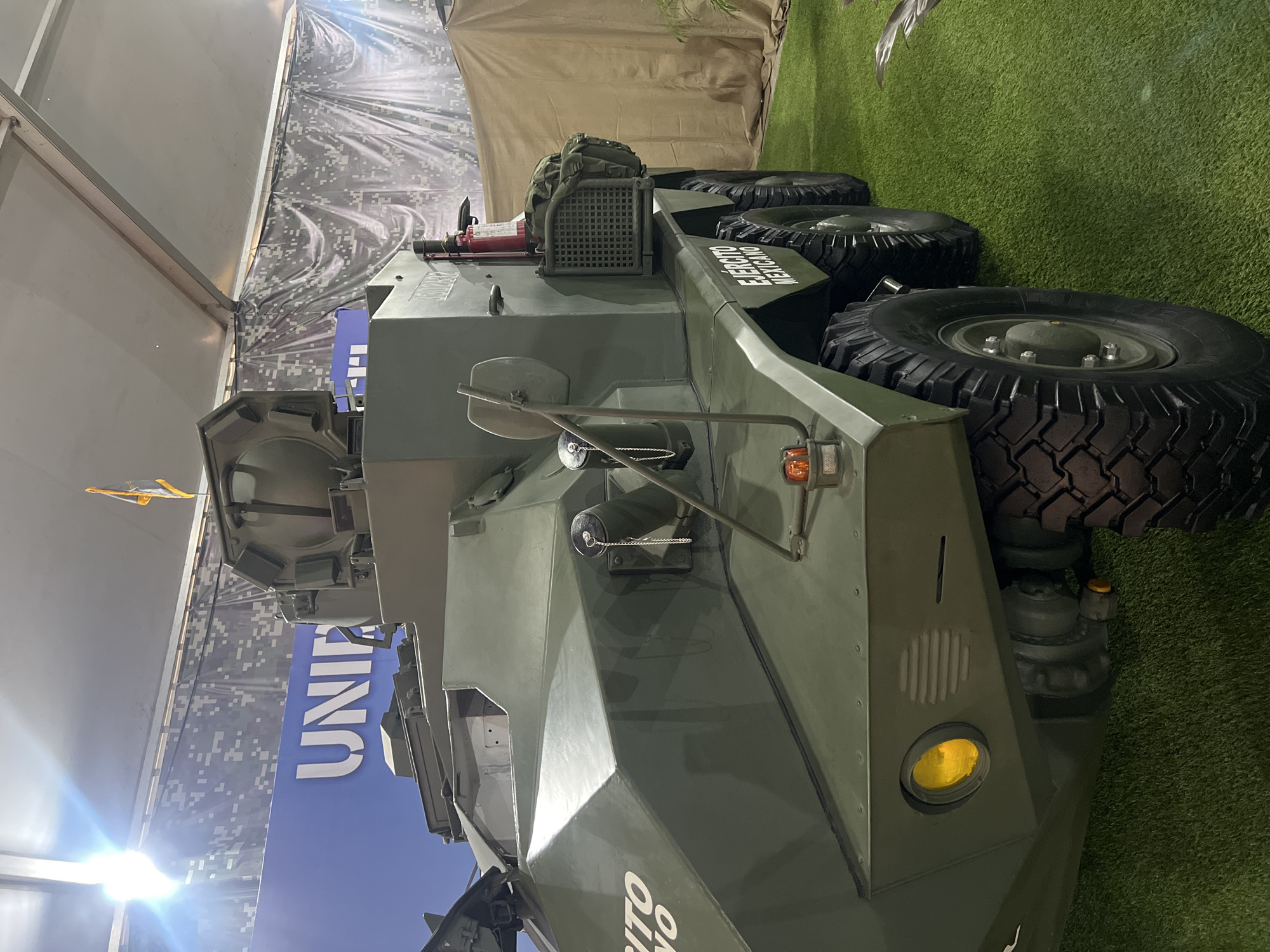 The image shows a close-up view of a military vehicle, likely an armored personnel carrier or similar type of vehicle. The vehicle is painted in a matte olive-green color and appears to be positioned indoors, possibly at a defense expo or exhibition. The vehicle has large tires and a boxy, armored design. The words "EJERCITO MEXICANO" are visible, indicating that it belongs to the Mexican Army. The background includes a blue sign with the word "UNIF" on it and a camouflage patterned backdrop. The floor is covered with artificial green turf.