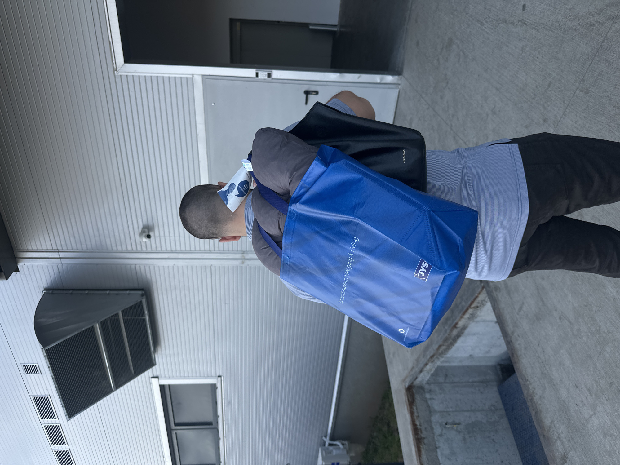 The image, oriented as if upright, shows a person viewed from behind, walking or standing outdoors next to a building. The individual is a male with very short, shaved hair, wearing a light blue/grey striped shirt and dark grey/black pants. He carries two bags: a large blue reusable shopping bag slung over his left shoulder and a black shoulder bag or tote over his right shoulder. A small white paper is stuck to the back of his neck/head area.
The background features the exterior of a building with light grey, horizontally corrugated metal paneling. Visible elements on the building include several vents, one large industrial-style exhaust vent, and a window. A dome-shaped security camera is mounted on the facade. A grey metal door with a handle is positioned near the person. The ground is concrete, and a concrete ledge or step is present on the right side of the frame.
Notable details include the text on the blue shopping bag, which reads "Scandinavian Sleeping & Living" and displays the "JYS" logo, along with a recycling symbol. The black bag shows the brand name "COCCINELLE". The white paper on the person's neck has an illustration of a man's face with a speech bubble containing partial text, including phrases like "...more good", "...on people", and "...opportunity". The scene is brightly lit by natural daylight, suggesting clear weather. The setting appears to be an urban or commercial area, consistent with locations in Sofia, Bulgaria.