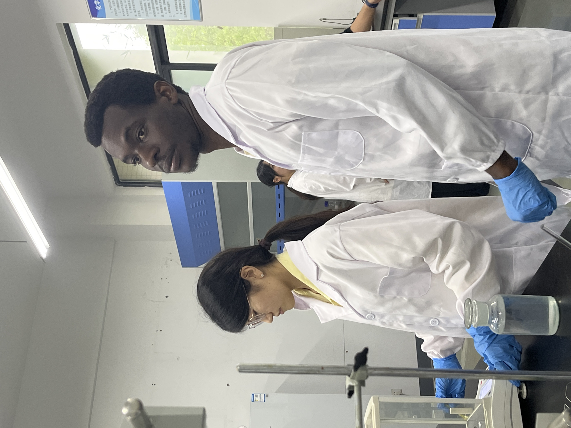 In a laboratory setting, two individuals wearing white lab coats and blue gloves are visible. The individual in the foreground, an Asian female, has dark hair pulled back in a ponytail and wears glasses. She is focused on a laboratory setup that includes glassware and support equipment. To her right, another individual, a Black male, is seen observing the experiment. The backdrop includes laboratory equipment, cabinets, and a sign with Chinese characters, suggesting a location in China. Based on the description and the location, it's possible that this image was taken at a laboratory in Jiangjin, China, as requested.