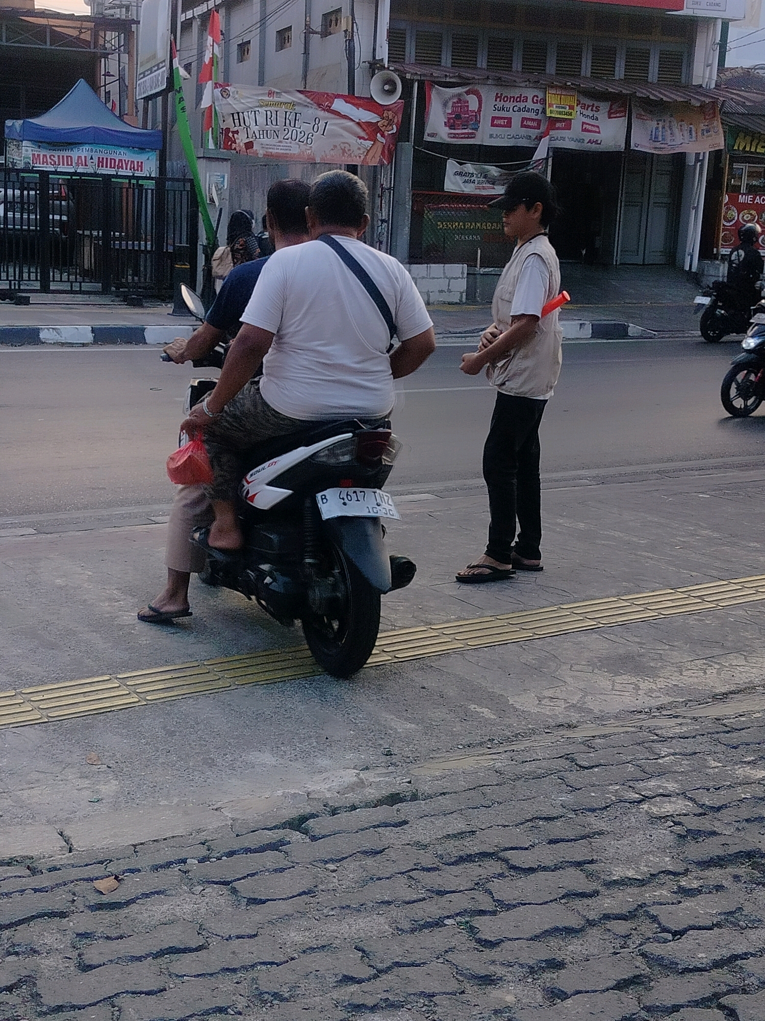 The image depicts a street scene in East Jakarta, Indonesia, likely near a commercial area given the storefronts and advertisements visible. In the foreground, two individuals are on a motorcycle. The rider, a male, is wearing a white t-shirt and patterned pants, with a red plastic bag tied to the back of the motorcycle. The passenger, also a male, is wearing a white t-shirt and camouflage-patterned shorts. They are stopped at what appears to be an intersection or a pedestrian crossing marked by textured yellow tactile paving.
To the right of the motorcycle, a person wearing a light-colored vest over a white t-shirt and black pants is standing on the sidewalk. This individual has an orange object attached to the back of their vest. Behind them, a storefront with various banners and signs is visible, including one that reads "Honda Gemilang Parts" and another with "MIZ..." suggesting a food establishment. Further in the background, more buildings and a blue tent with text like "MASJID AL HIDAYAH" are present. The road surface is asphalt and shows signs of wear and cracking. Other motorcycles are visible further down the street. The overall scene suggests a typical urban environment with daily activities taking place.