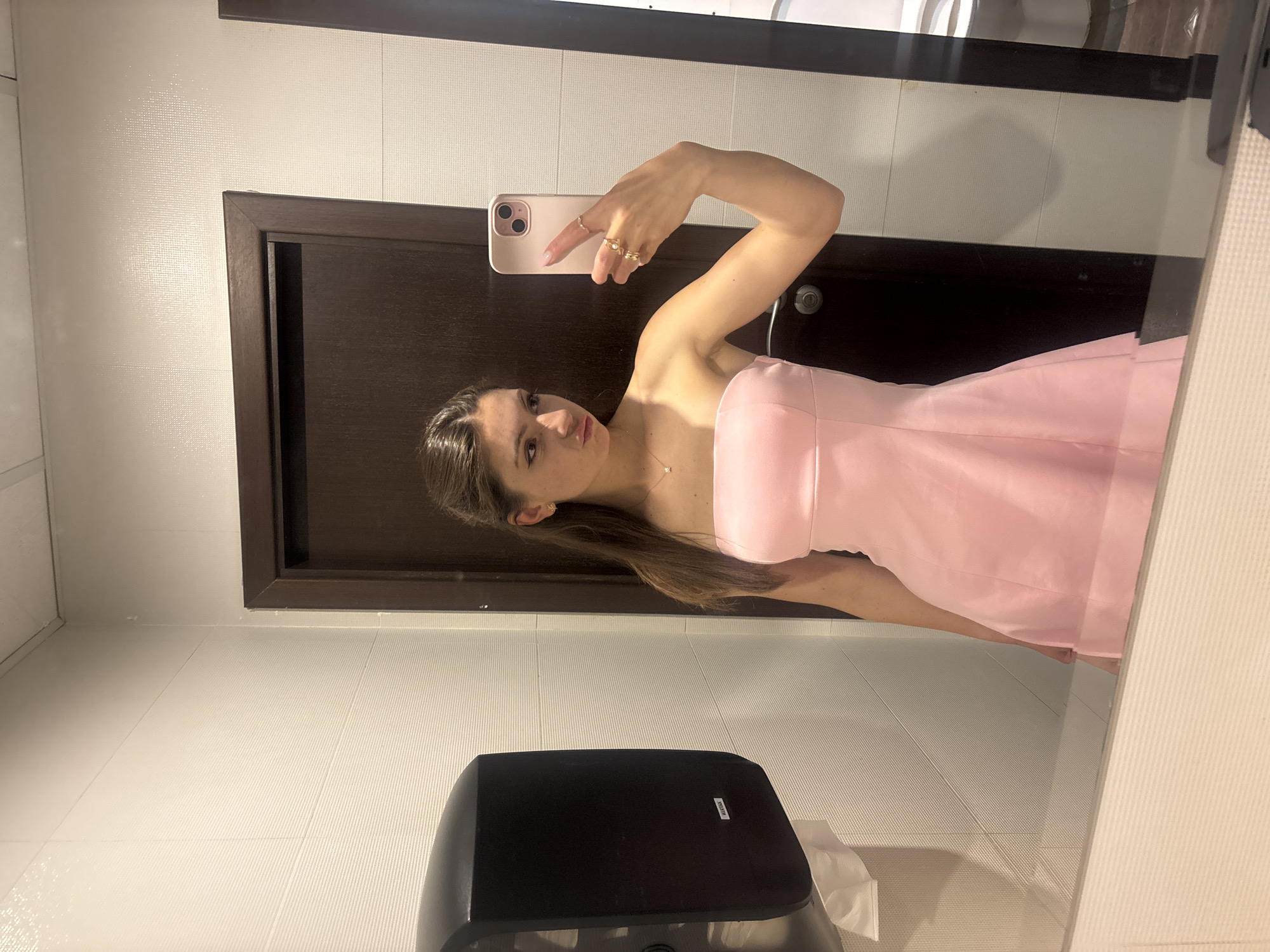 A woman in a strapless pink dress is taking a selfie in what appears to be a public restroom. She is holding a light pink iPhone. The background includes a dark brown door, white tiled walls, and a black automatic hand dryer. A tissue dispenser is partially visible. The location cannot be determined as Shumen, Bulgaria.