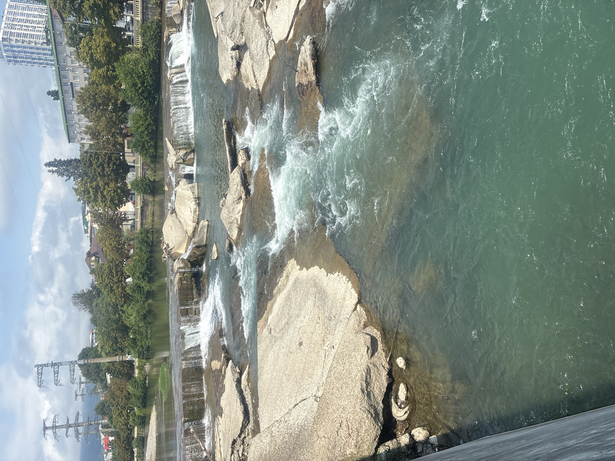 The image shows a view of the Matsesta River in Sochi, Russia, on a cloudy day. The river flows over a series of rocky ledges, creating small waterfalls. In the background, there are trees, buildings, and power lines. The water is a murky green color.