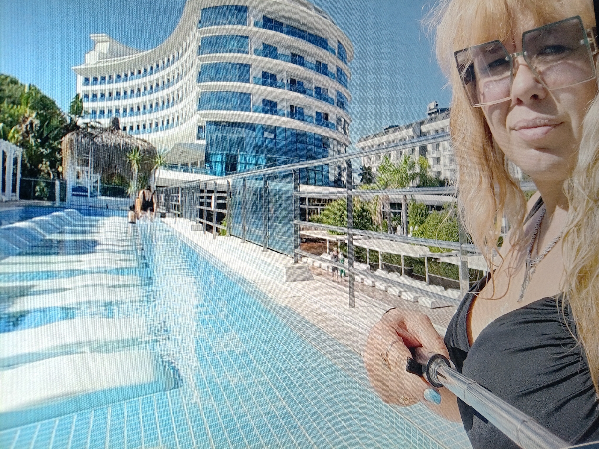 The image depicts a selfie taken at a resort. A woman with blonde hair and large square sunglasses is holding a selfie stick, partially obscuring her face. She is wearing a black top. In the background, a curved, multi-story white building with blue-tinted windows dominates the scene, suggesting a hotel or resort. A swimming pool with blue tiled steps fills the foreground. Lush green trees and other resort structures are visible. The environment appears to be sunny with a clear blue sky. The image does not contain any elements that would suggest the location is the city of Tiraspol, Moldova.
