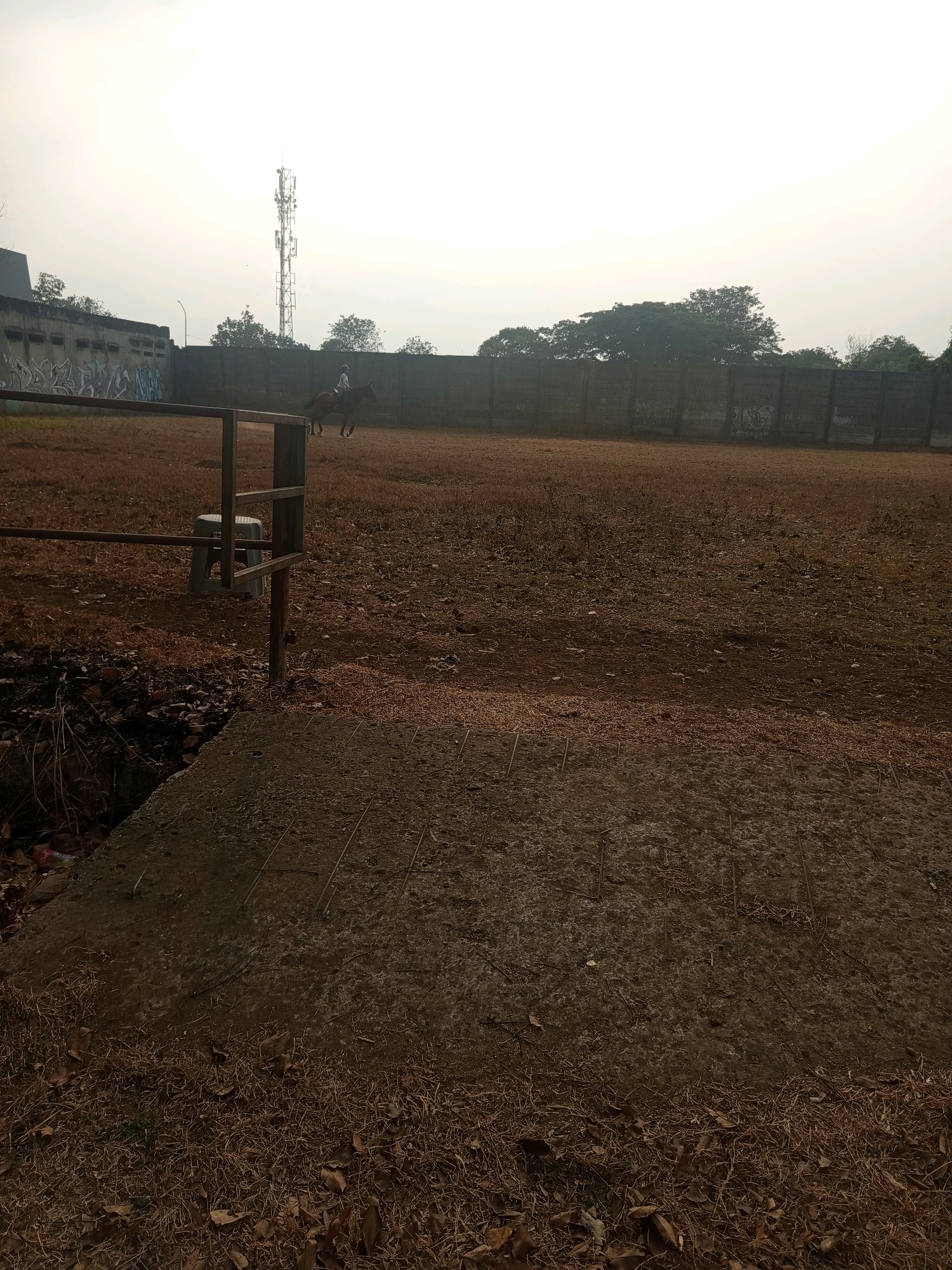 The image shows an outdoor equestrian arena in West Jakarta, Indonesia. A person is riding a brown horse in the center of the dusty arena. A tall telecommunications tower is visible in the background, along with a concrete wall covered in graffiti. The ground is covered in dry grass and leaves, suggesting it may be a dry season. A metal fence and a small plastic stool are in the foreground, near a concrete slab with exposed rebar. The sky is overcast, giving the scene a muted and slightly hazy appearance.