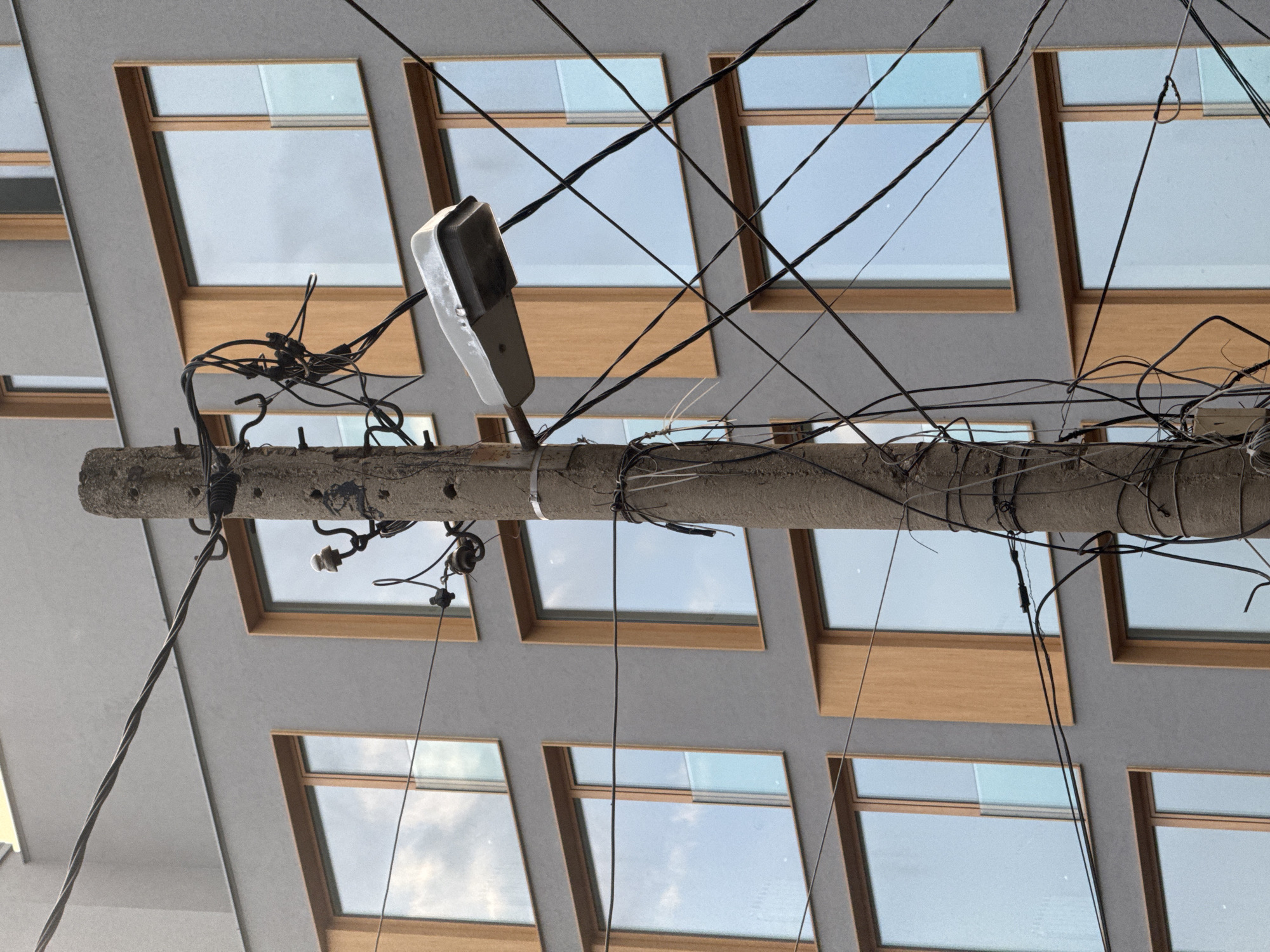 The image shows a concrete utility pole covered in numerous electrical wires and a street lamp. The pole is situated in front of a modern building with a gray facade and rectangular windows framed in wood. The building appears to be in an urban setting. The wires are messy and tangled, draped across the frame and obscuring parts of the building. The street lamp is mounted on the pole and is facing towards the right of the frame. The sky is visible through the windows, reflecting a cloudy, possibly overcast day. The scene is static, with no people or animals visible.