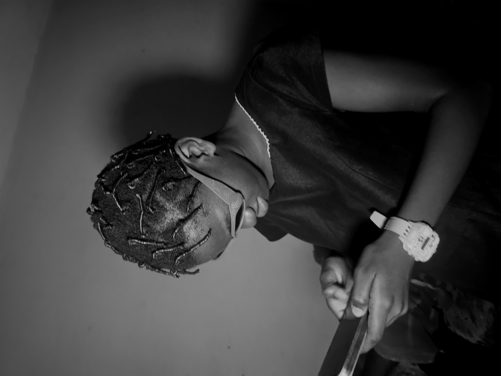 A person is holding a knife in Nkwelle, Nigeria. The individual is dark-skinned, wearing a dark-colored top, and a light-colored digital wristwatch on their left wrist. They have a short hairstyle with small braids. The person appears to be looking down, holding a knife in their right hand. The background is a plain wall. The image is in black and white.