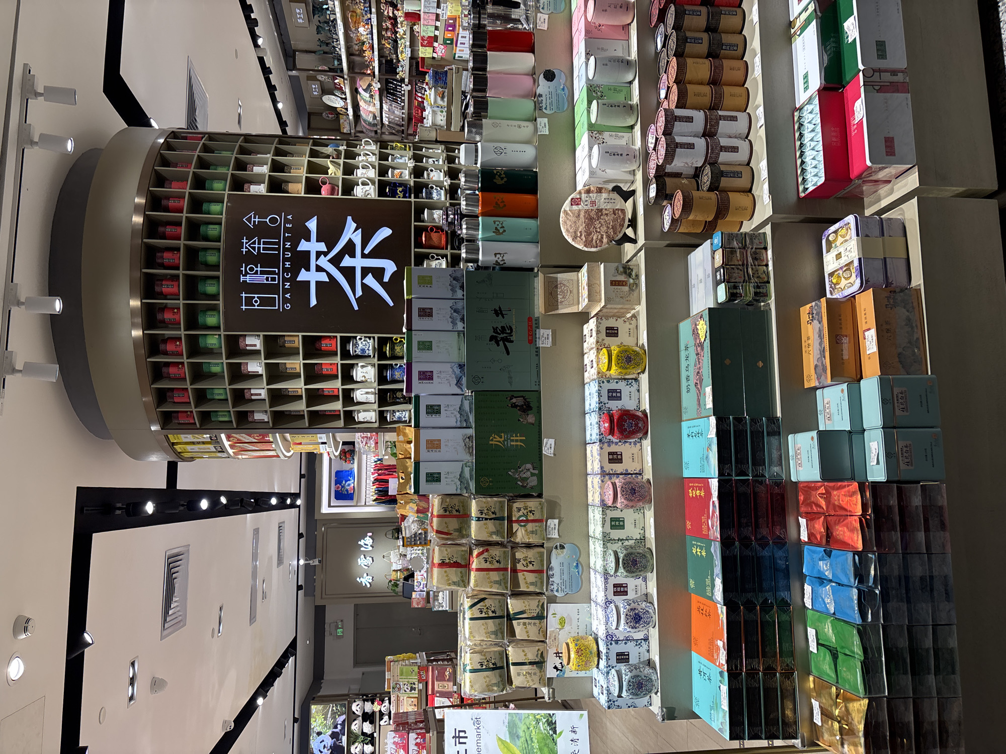 This appears to be a photograph taken inside a tea shop, likely located in Guangzhou City, China. The store seems well-stocked with a variety of tea products, organized on shelves and displays. The tea is presented in different types of packaging, including boxes and cylindrical containers of various colors and designs. There is also merchandise such as teacups and other teaware on display. A sign with Chinese characters and the English name "GANCHUNTEA" is visible. There are also signs promoting types of tea, such as "Long Jing" and "Tieguanyin." The lighting is bright. The image is taken from a relatively high angle, possibly from near the ceiling or an upper level of the store.