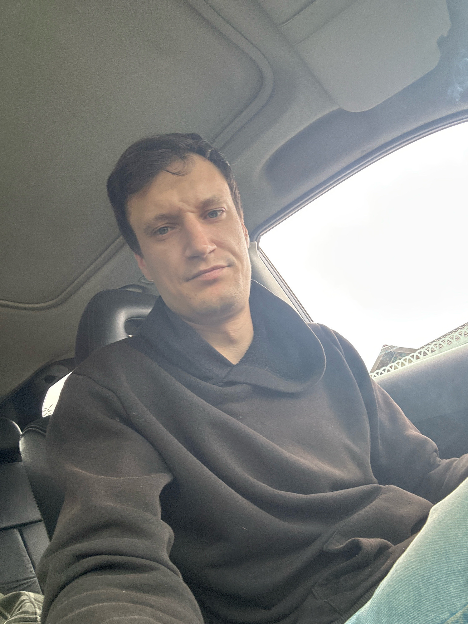 A Caucasian male in his late 20s or early 30s is in the driver's seat of a car, looking directly at the camera. He has short, dark brown hair and fair skin. He is wearing a dark brown hooded sweatshirt and light blue jeans. The interior of the car is visible, including the black leather headrest of the passenger seat and the grey headliner. The sunroof is open, and the sky is overcast. In the background, a building with an ornate white railing is visible through the passenger window. The image was taken in Makiv, Ukraine.