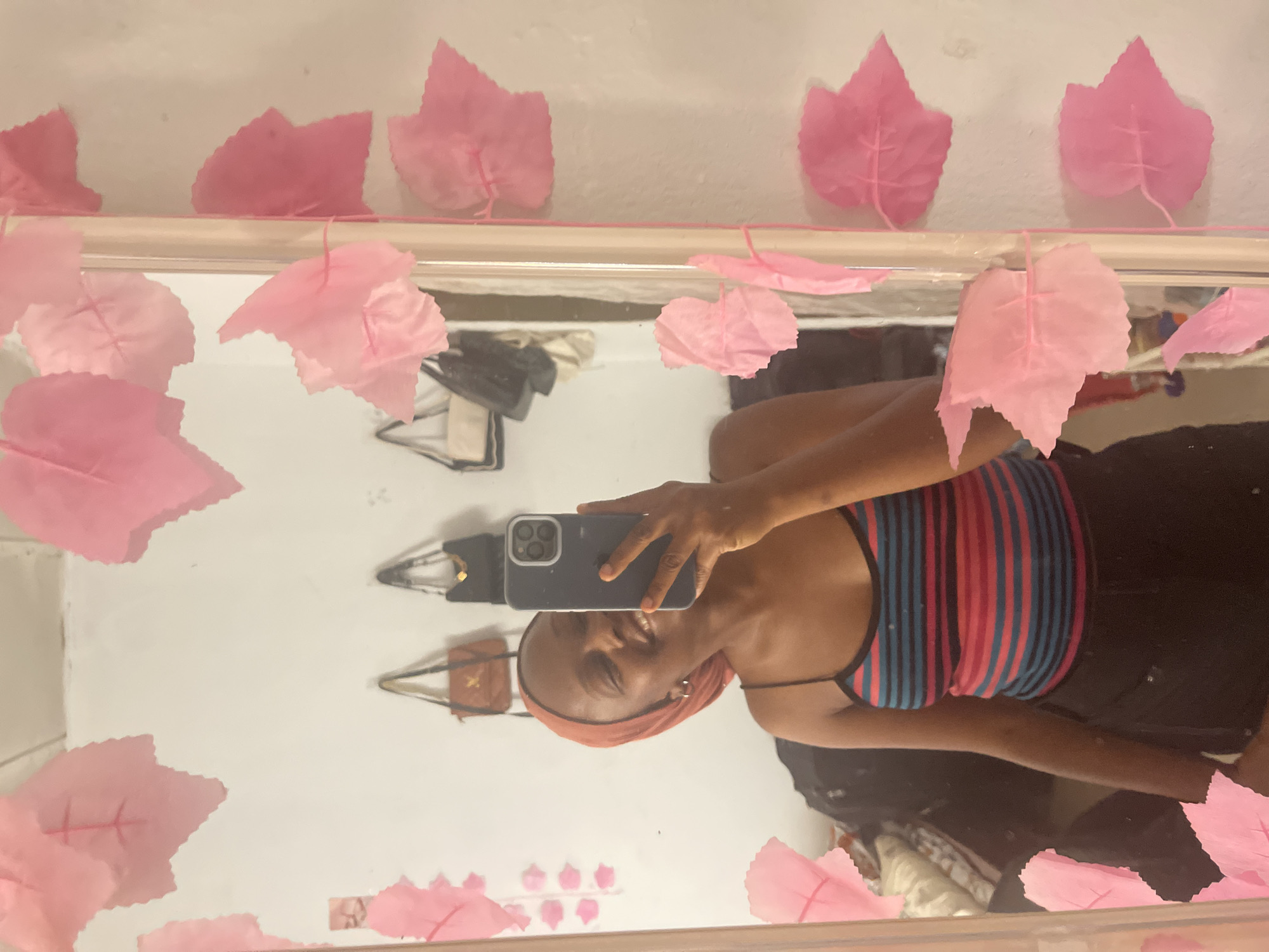 The image shows a young woman taking a mirror selfie in a room. She is wearing a striped tank top and black shorts. She has a headscarf on and is smiling. The mirror is decorated with pink paper leaves. In the background, several handbags are hanging on the wall. The overall impression is of a casual, personal space. The image location is stated to be Jos, Nigeria.
