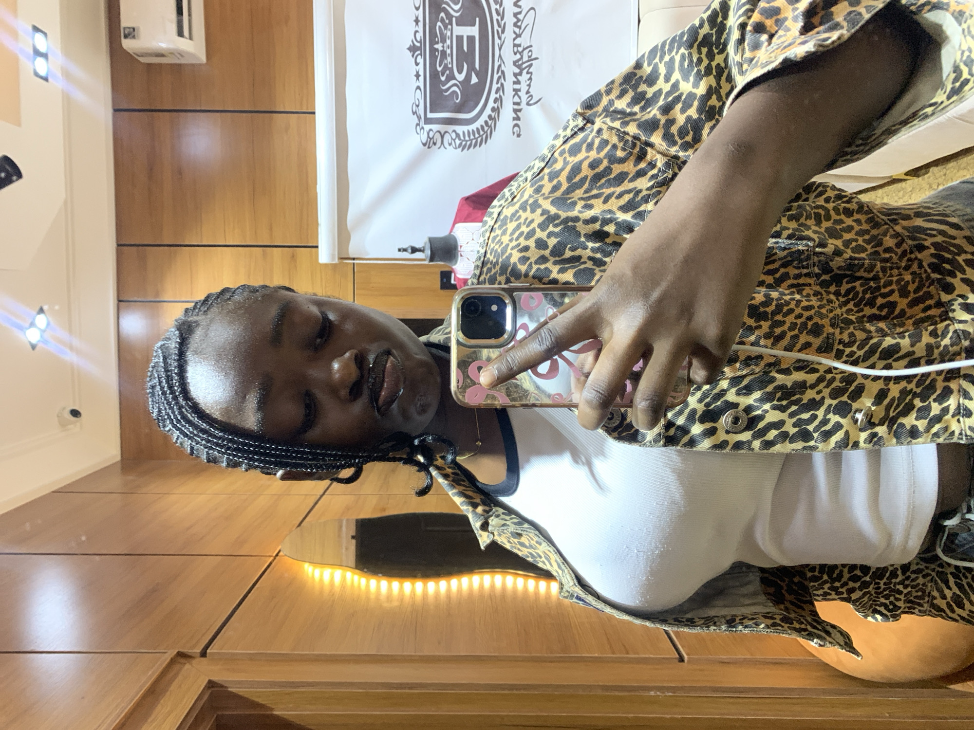 A young Black woman with braided hair takes a mirror selfie in a room. She is wearing a white ribbed tank top and a leopard print denim jacket. Her lips are pursed, and she is holding a phone with a pink case in her right hand. The background shows wood-paneled walls, a security camera, an air conditioning unit, and a white banner with a crest and text. The setting appears to be an indoor boutique or store in Gura Top, Nigeria.