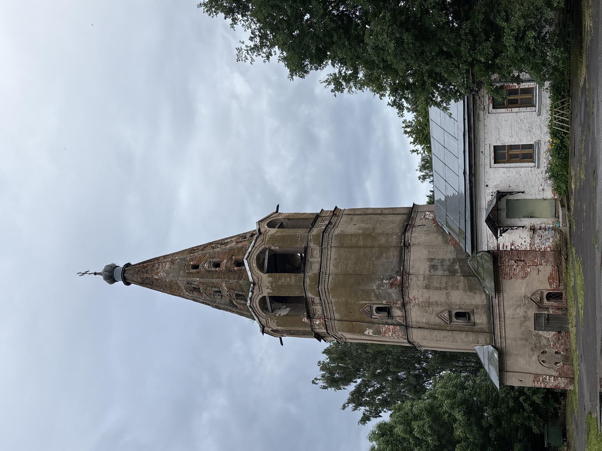 An aged, octagonal bell tower of a church stands prominently against an overcast sky. The tower, constructed of brick and plaster, shows significant signs of disrepair with crumbling sections revealing underlying red brick. It is topped with a dark, onion-shaped dome and a cross. Adjacent to the bell tower is a smaller, single-story white building with a silver, sloped roof and two wooden-framed windows. A green door with an awning is visible on the front of this building. Lush green trees surround the structures, and the ground in front is paved with asphalt, wet in places, suggesting recent rain. The overall scene depicts a weathered, historical religious site. This is the bell tower of the Church of the Ascension of the Lord, located in Veliky Novgorod, Russia.