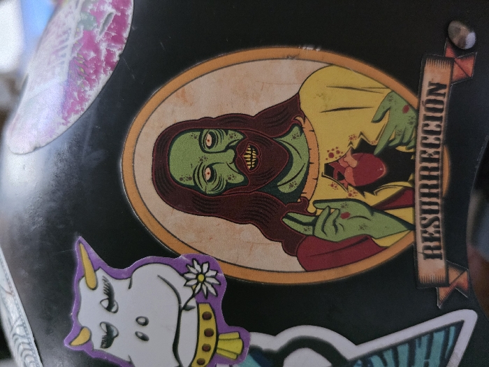 The image displays a close-up of multiple stickers applied to a curved, black surface, likely a helmet or similar object, exhibiting minor scratches and dust. The central and dominant sticker is an oval-framed illustration of a figure resembling Jesus Christ, but depicted with zombie characteristics. This figure has green skin, dark red hair and beard, pale yellow eyes with dark pupils, and an open mouth revealing sharp, exposed teeth. Bloodstains are visible on the figure's face and hands. The figure wears a yellow tunic and a red cloak. Its left hand is raised with two fingers extended, showing a red mark (stigmata) with dripping blood. The right hand points to an anatomical heart, rendered in red, visibly exposed through a jagged opening in the figure's chest, surrounded by red splatters. Below this oval sticker, a banner with faded brown shading reads "RESURRECCIÓN" (Spanish for "RESURRECTION") in a bold, distressed font. To the top-left of the zombie Jesus sticker, a cartoon sticker of a white cow is visible. This cow has purple horns and outline, closed eyes with long eyelashes, a small smile, and holds a white flower with a yellow center in its mouth area, while wearing a yellow and brown collar. A partially visible, faded sticker with pink and white patterns is located in the top-right corner of the black surface. The specific location in the city of Villa San Andrés, Argentina, cannot be determined from the visual information provided in the image.