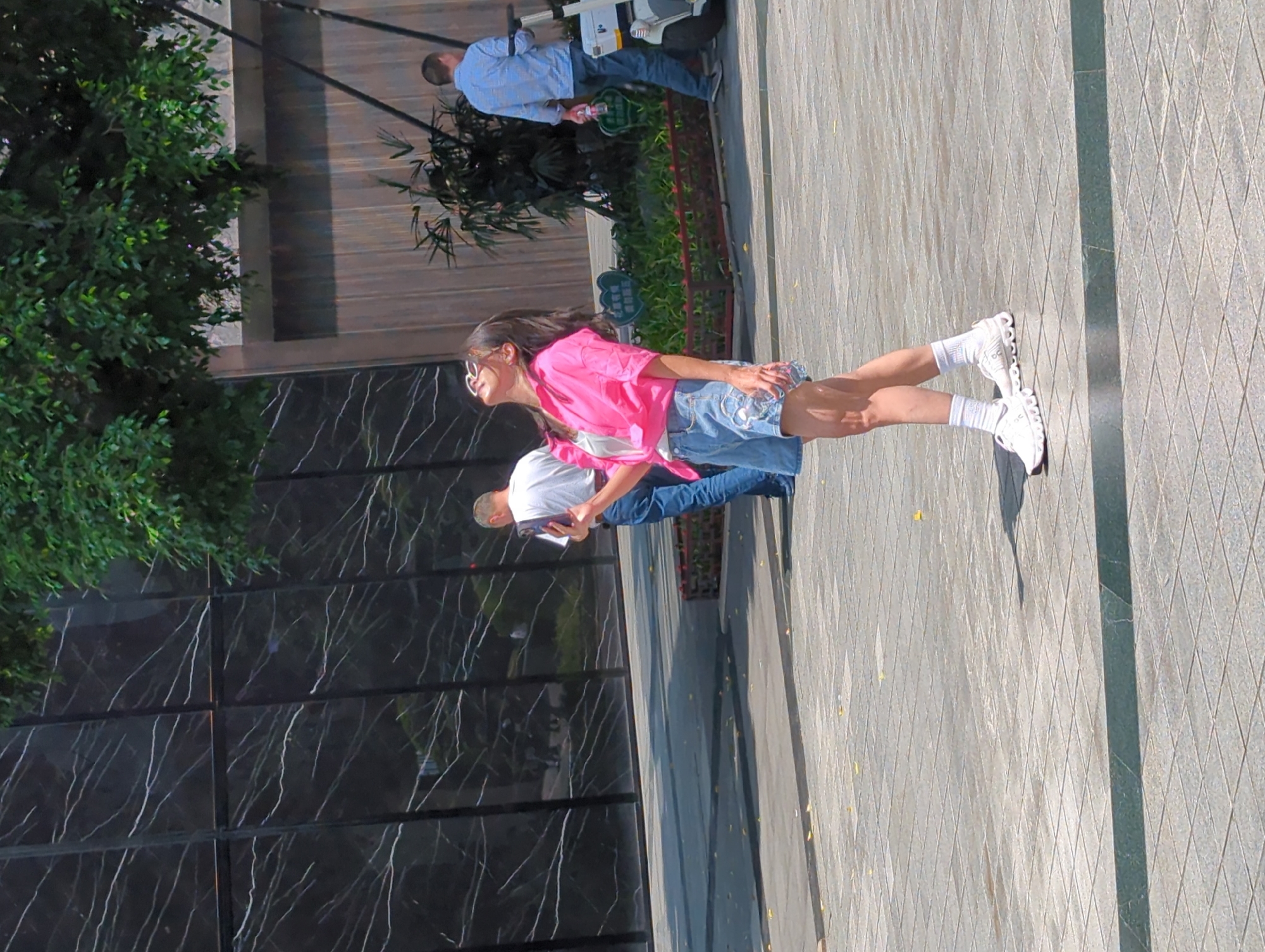 On a sunny day in Yuzhong District, China, a young woman stands on a paved public space, wearing a pink shirt, jean shorts, and white roller skates. She looks off to the side. Nearby, a man in a white shirt holds a notebook. In the background, a glass-paneled building is partially obscured by a large green tree. Further in the distance, another man in a blue plaid shirt is pushing a cleaning device. The setting is outdoors on a textured, grey paved surface.