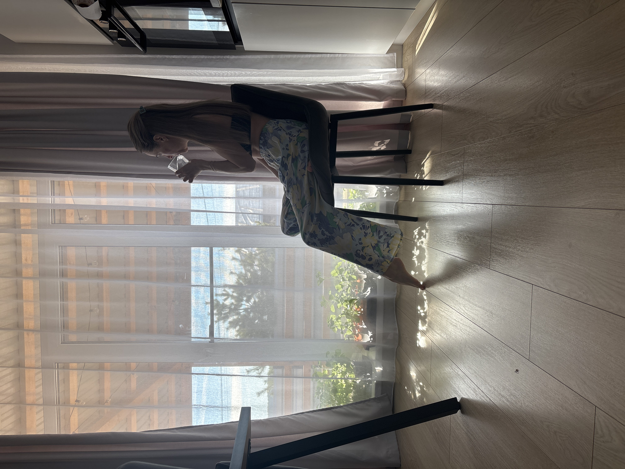 A woman is depicted sitting on a black chair, leaning forward, and holding a clear glass or cup. She has long hair, secured with a clip, and is wearing a dark top and floral-patterned wide-leg pants. Her bare feet are visible on the light-colored wooden floor.
The scene is an indoor setting, brightly lit by natural sunlight streaming through a large window that spans most of the background. The window is covered with sheer white curtains, with darker grey curtains pulled to the sides. Outside the window, a glimpse of trees and possibly another building can be seen, along with potted plants on the windowsill. Above the woman, part of a kitchen area is visible, including white cabinets and a built-in oven or microwave. The sunlight casts distinct patterns and shadows across the wooden floor, indicating a sunny daytime. The overall impression is a modern, light-filled home environment. There is no visible text in the image.