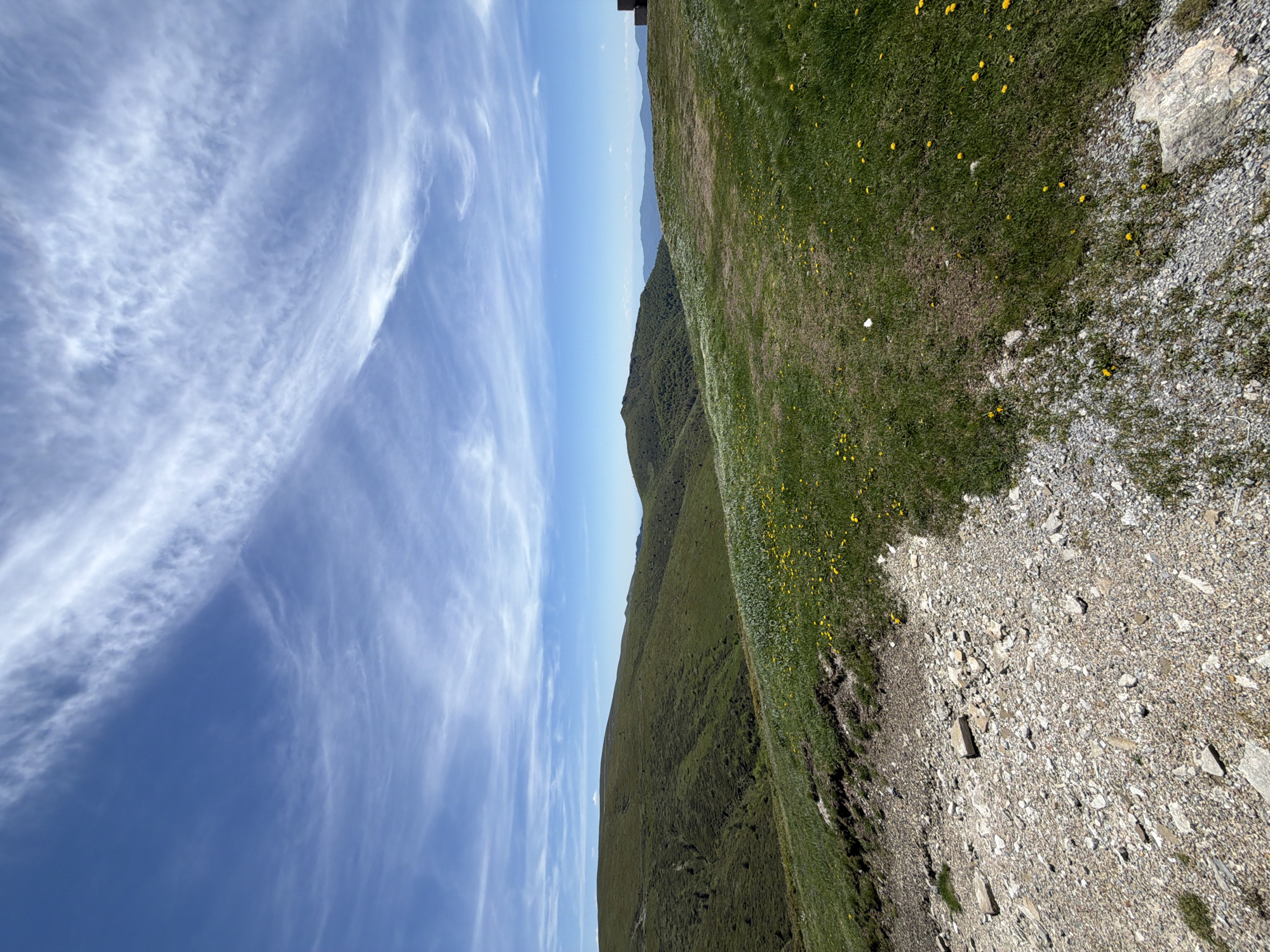 The image shows a landscape in Beli Osam, Bulgaria. The sky, filled with wispy, streaky clouds, occupies the left side of the frame. To the right, a series of rolling green hills stretch toward the horizon. The foreground contains a mix of grassy areas dotted with small yellow flowers and a section of rocky, gravelly ground.