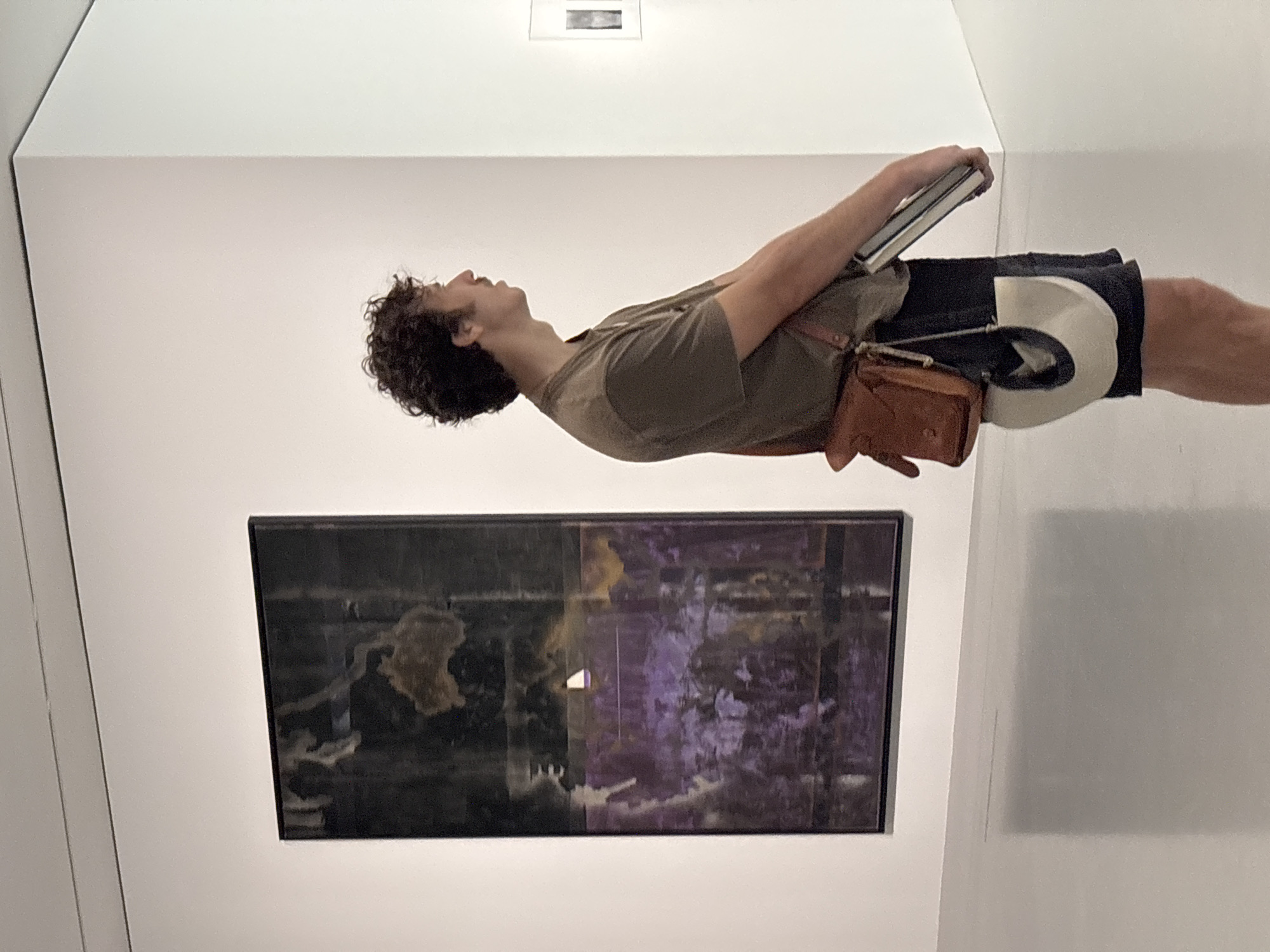 The image depicts a young man with curly dark hair and a mustache standing in a well-lit, minimalist art gallery. He is wearing a short-sleeved olive green t-shirt, dark shorts, and a brown leather shoulder bag. He is holding a book or portfolio in his right arm, and his left hand is resting on the book.
To his left, on a white wall, hangs a large, abstract painting. The painting is framed in black and features dark, deep purples and blacks with lighter, abstract shapes and textures. There is a small, rectangular bright white element near the center of the painting.
To the man's right, on another white wall, is a smaller framed artwork, possibly a photograph, with a dark frame and a light background. The room has polished concrete or stone floors and bright, diffused lighting from an unseen source. The overall impression is one of modern, clean aesthetics. The scene is set in Paris, France.