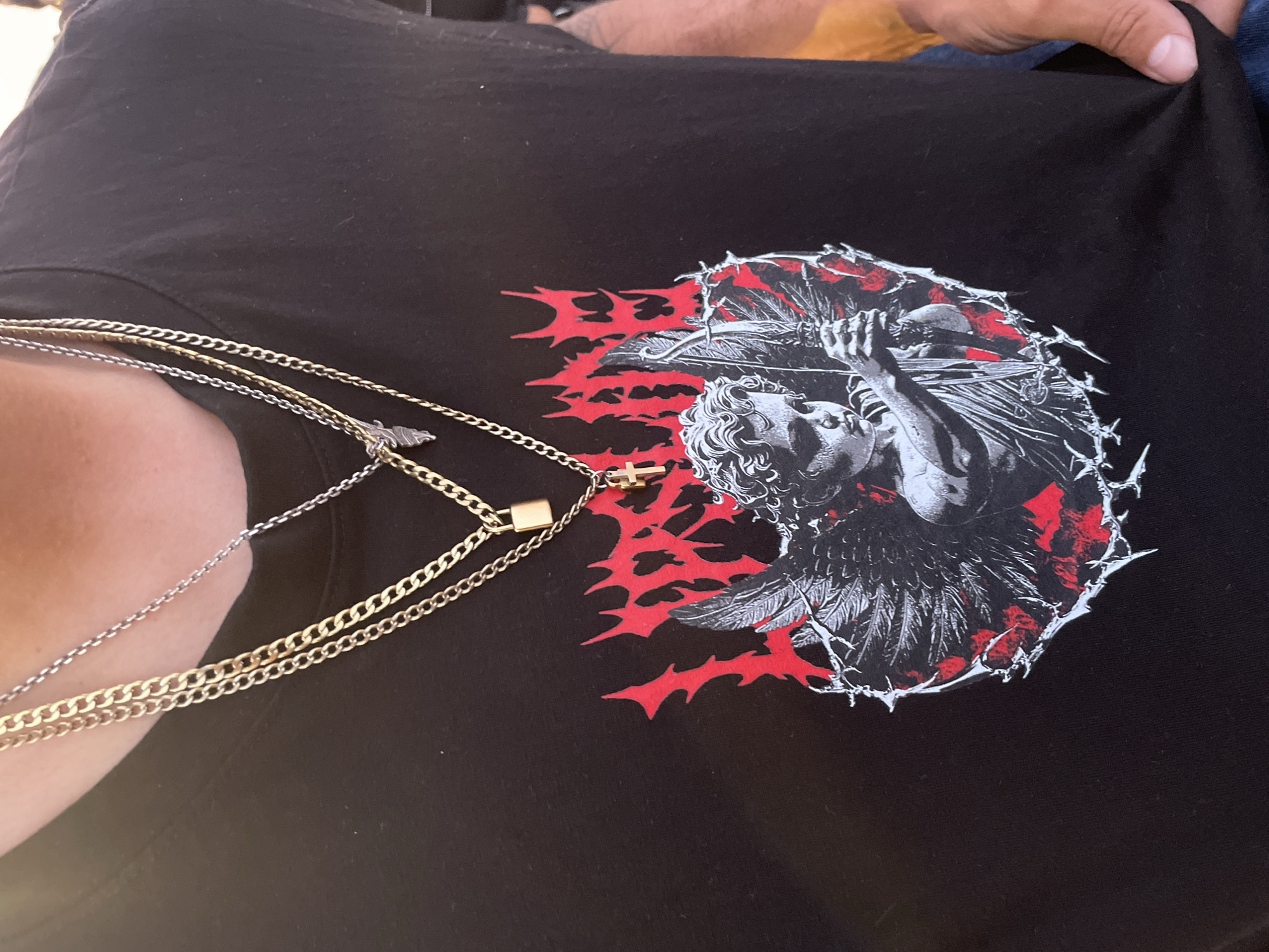 The image shows a close-up of a person wearing a black t-shirt with a graphic print. The print features an image of an angel-like figure with wings, surrounded by red and white patterns. The person is also wearing multiple gold and silver necklaces. The background is blurry and indistinct, making it impossible to determine the location. Therefore, it is not possible to confirm if the image was taken in Barcelona, Spain.