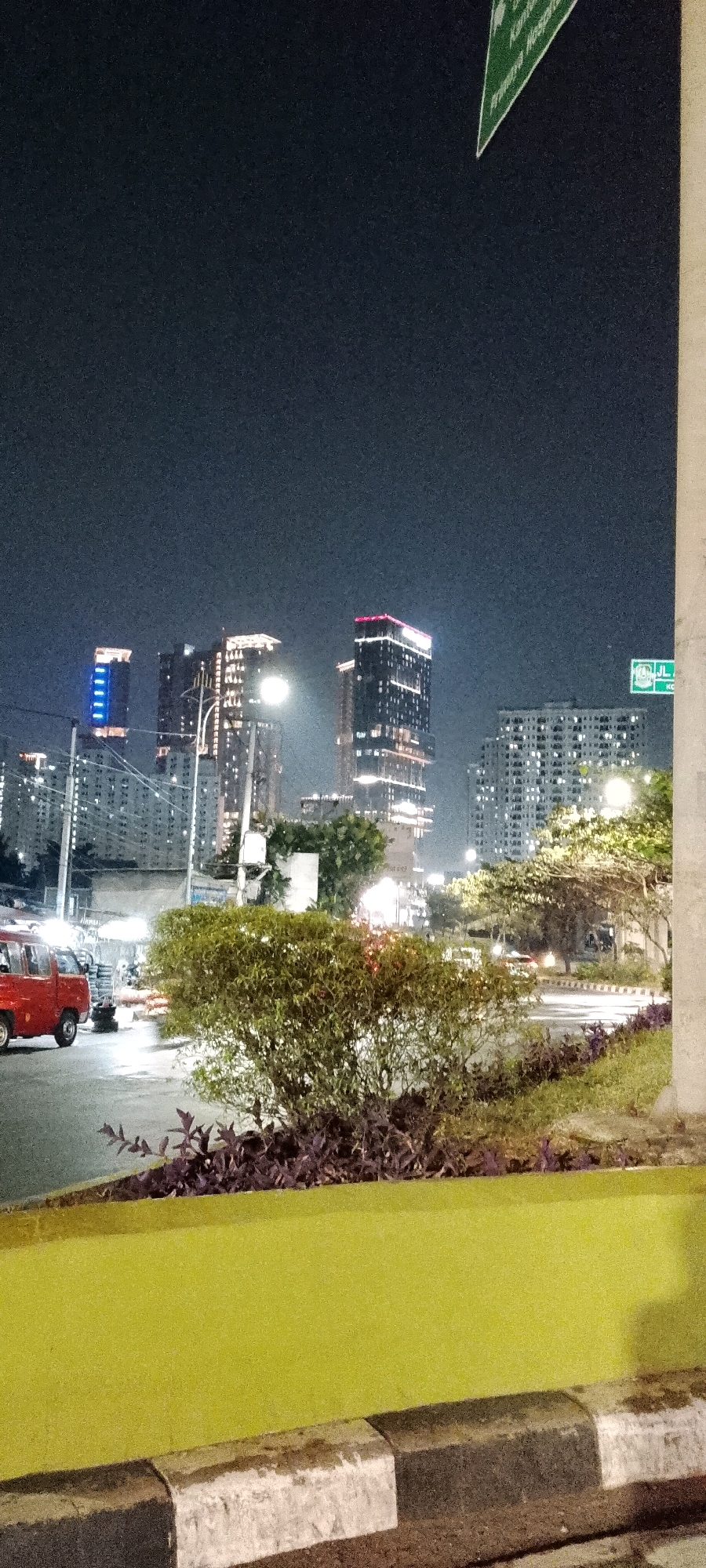 A nighttime photograph captures a cityscape in Bekasi, Indonesia, featuring illuminated skyscrapers against a dark sky. In the foreground, a bright yellow concrete barrier with a gray and black curb runs horizontally across the bottom of the frame. Behind the barrier, a lush green bush is prominently featured, with clusters of purple-leafed plants growing beneath it. To the left of the bush, a red vehicle is parked on a road. The middle ground shows more greenery, trees, and streetlights. The background is dominated by several tall buildings, their windows and decorative lighting casting a warm glow. A signpost with green lettering is visible in the upper right corner of the image. The overall scene suggests a well-lit urban environment at night.
