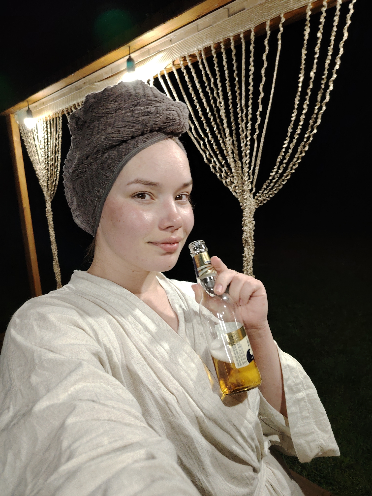 A young woman, likely after showering, stands outdoors at night, possibly in the city of Malyi Bukor, Russia. She is wearing a light-colored robe and has a grey towel wrapped around her head. In her right hand, she holds a bottle of beer, partially consumed. Behind her, there is a wooden structure with strings of what appears to be decorative lights. The background is dark, with some faint green grass visible near the bottom right corner. The lighting suggests an artificial source, possibly from the structure.