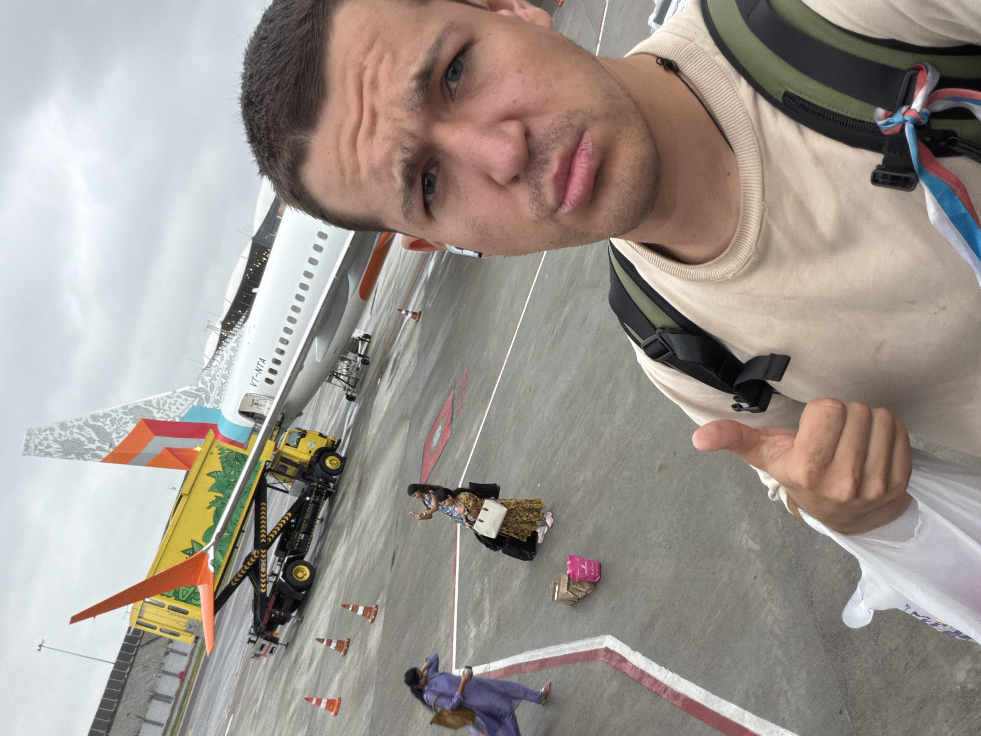 The image shows a man taking a selfie on an airport tarmac. He is giving a thumbs-up gesture and has a playful expression. Behind him, an airplane is parked, with baggage handling equipment and other airport personnel visible. The weather appears to be overcast. The image does not provide any specific information that would allow for its localization in Hyderabad, India.
