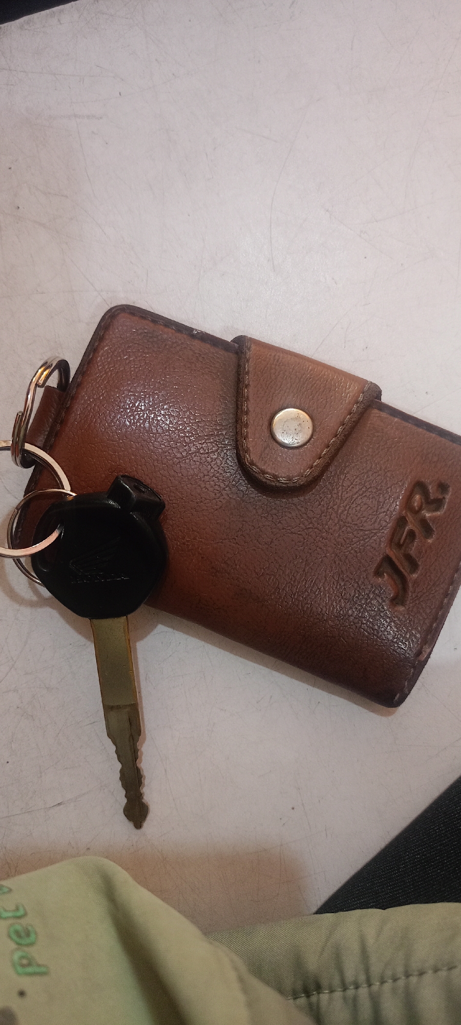 A brown leather key holder with a metal snap closure is on a scratched white surface. A metal keychain ring with a black key fob and a single brass key is attached to the key holder. The key fob has the word "HONDA" embossed on it. The letters "JFR" are embossed on the key holder. A light green fabric with green lettering is partially visible in the bottom left corner. The image does not contain enough information to determine its location within the city of Mekar Rahayu, Indonesia.