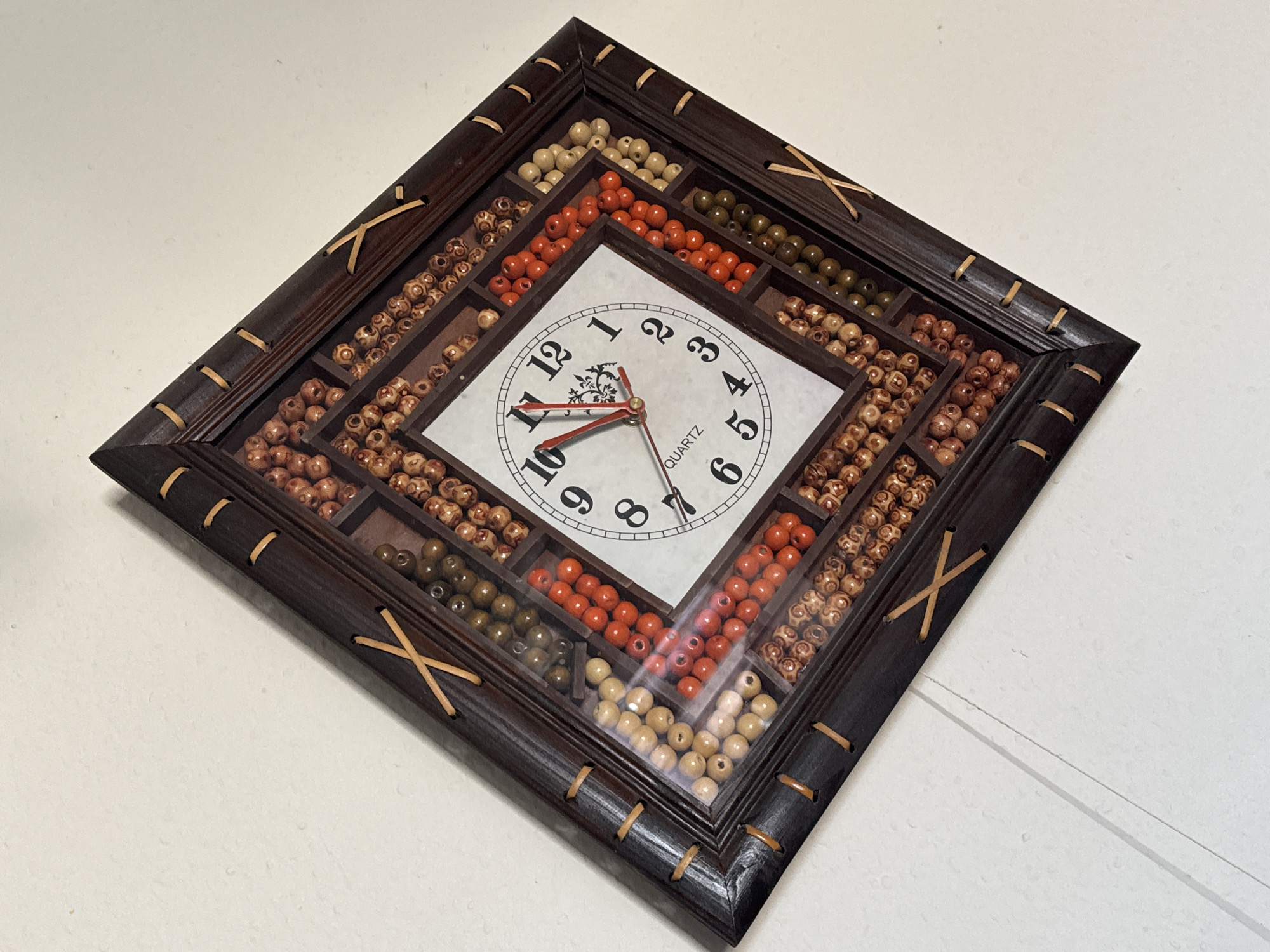 This image shows a close-up, angled shot of a decorative wall clock against a plain white wall. The clock has a dark wooden frame with a diamond pattern formed by small wooden sticks crisscrossing at the corners and along the edges. The clock face is square and white, with black Arabic numerals indicating the hours and black hands. The clock is surrounded by a grid of small compartments filled with various colored beads. These beads are arranged in rows and include shades of orange, tan, olive green, and light brown. The clock is tilted, with the top right corner pointing upwards and slightly towards the viewer. The background is a plain, textured white wall. There are no people or animals visible in the image. The lighting appears to be soft and diffused, casting minimal shadows. The image does not provide any information about its location in Bishkek City, Kyrgyzstan.