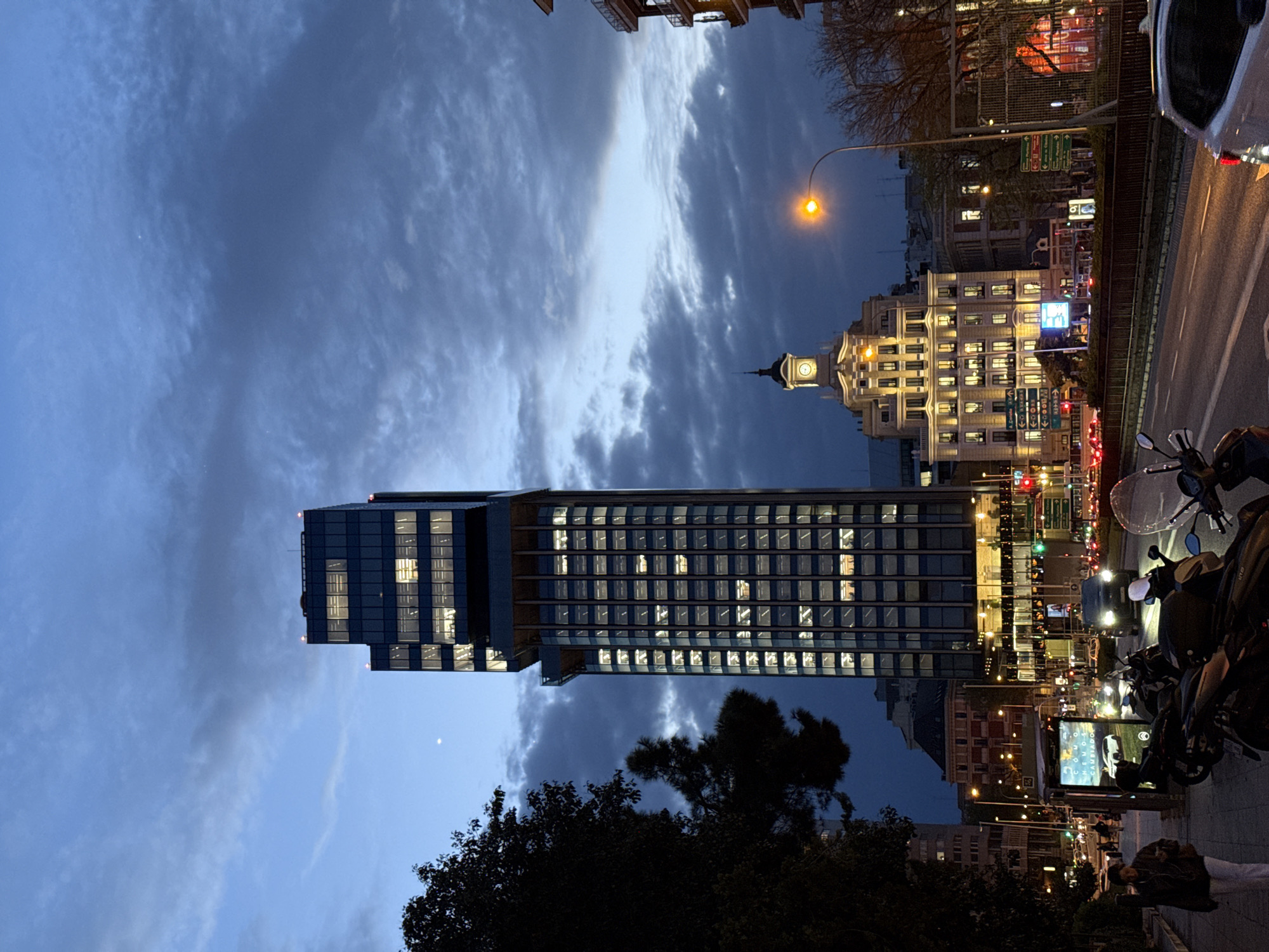 The image depicts an urban scene in Madrid, Spain, at dusk. A tall, modern skyscraper with illuminated windows dominates the view. To the right of the skyscraper is a traditional-style building with a clock tower, also illuminated. Streetlights cast an orange glow on the scene. The sky above is a mix of dark and light blue, with clouds scattered throughout. A busy street runs along the right side of the image with cars and motorbikes parked on the sidewalk. Silhouetted trees are visible at the bottom. A pedestrian can be seen walking on the sidewalk.