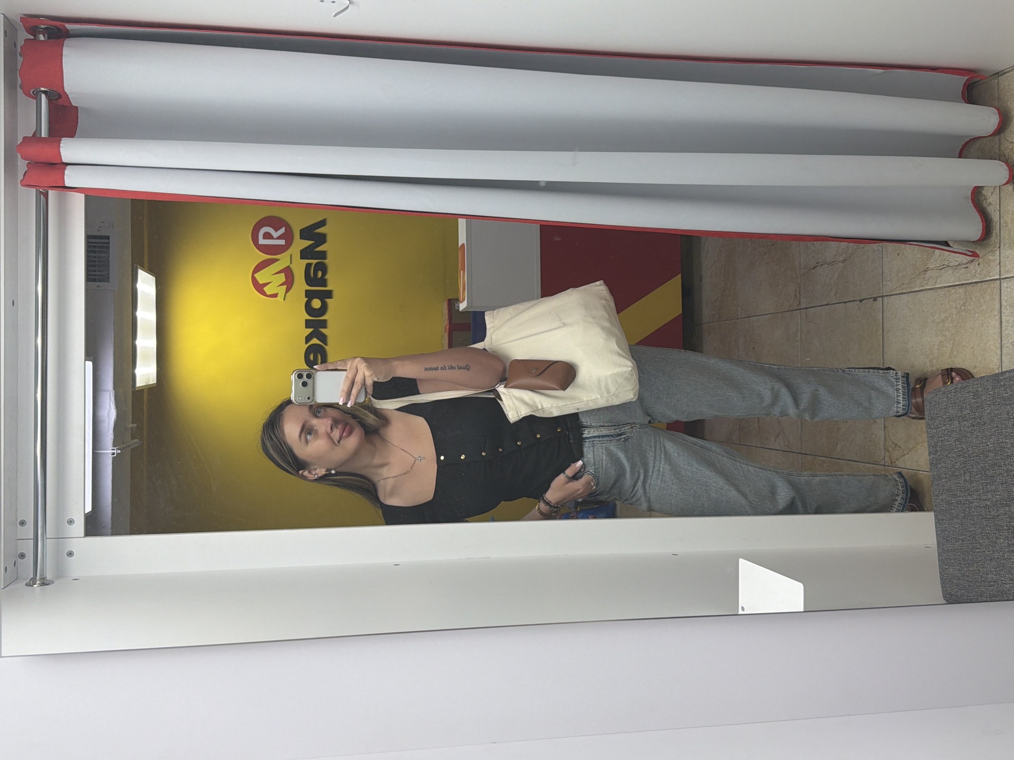 A female subject is captured in a full-length mirror selfie, positioned within a white-walled fitting room or changing area. The subject is a young adult with light-toned skin and long, light brown hair styled with a center part. She is wearing a short-sleeved, black square-neck top with several small gold buttons down the front, light blue high-waisted denim jeans with side slits at the ankles, and brown sandals. Accessories include a silver chain necklace with a cross pendant, multiple dark bracelets on her right wrist, rings on her fingers, and pearl drop earrings. She holds a silver or white smartphone with multiple camera lenses in her right hand, positioned to take the photo. A light-colored canvas tote bag with a small brown leather pouch attached to its strap hangs on her left shoulder. Textual script is tattooed on her left inner forearm.
The fitting room features a full-length mirror reflecting a retail environment. To the right of the mirror, a grey curtain with a red border and metal eyelets is partially drawn on a silver rod. To the left, a small white shelf is visible on the wall. A grey upholstered bench or stool is positioned in the foreground, partially out of frame and slightly out of focus. The floor is covered in light-colored square tiles.
The reflection in the mirror reveals a bright retail space with a prominent yellow wall. On this wall, a mirrored logo of two red circles containing the letters "M" and "Я" (Cyrillic for "R" or "Ya") is visible, with the mirrored Cyrillillic text "экрям" (which reads as "МАРКЕТ" or "MARKET" when unmirrored) below it. This indicates a retail market setting in a Russian-speaking country. Below the logo, a red counter or shelf is also reflected. Overhead, strip lighting and an air vent are visible in the reflection. Based on the Cyrillic text identifying it as a "MARKET" and the prompt's specification, the location is a retail market in the city of Moscow, Russia.