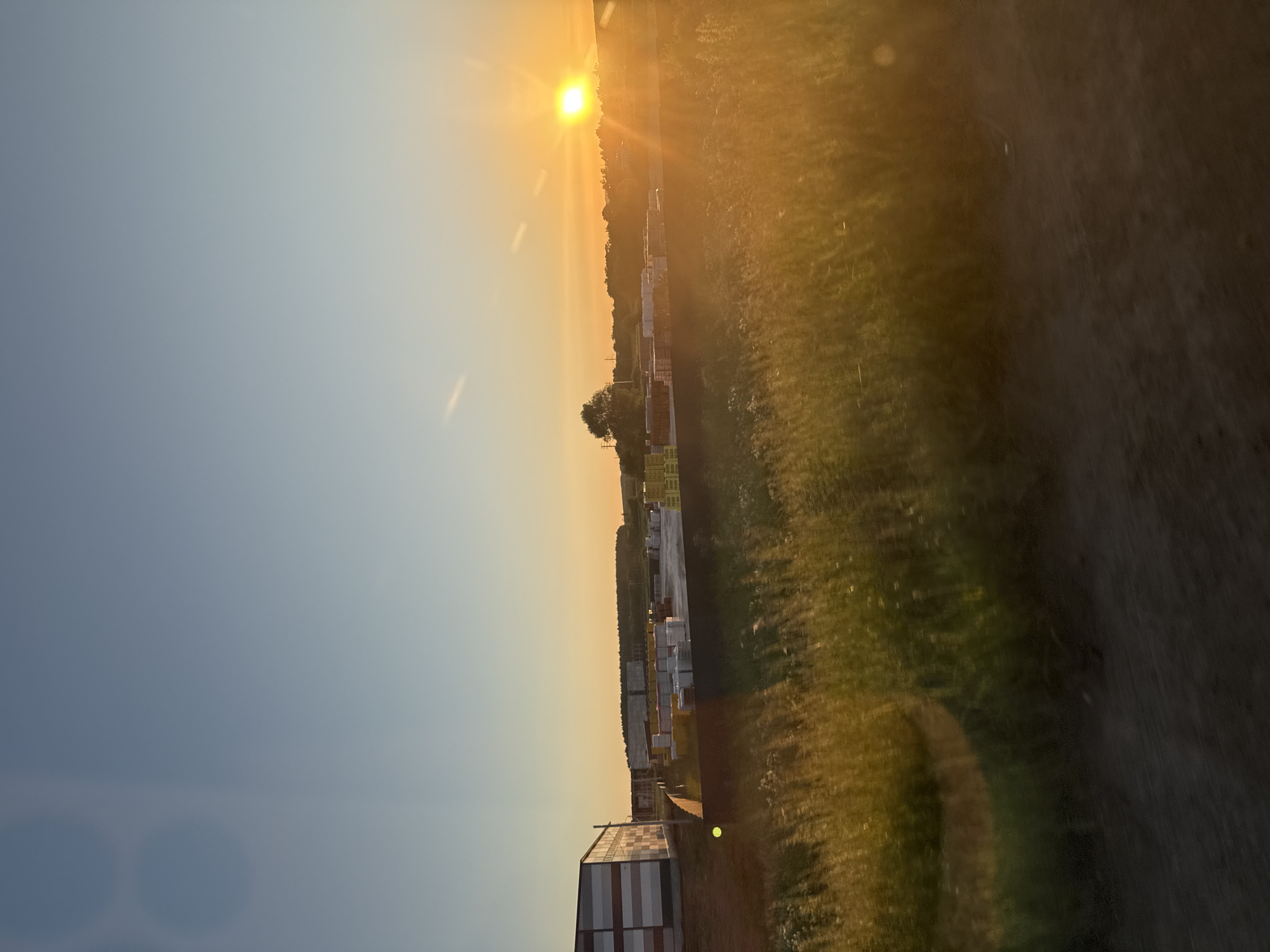 A wide shot captures an industrial area at sunset in Malaya Ven'ya, Russia. The sky transitions from a clear blue at the top to a warm orange near the horizon, illuminated by the setting sun on the right side of the frame. In the foreground, tall, blurred green grass is illuminated by the golden hour light, suggesting movement or a gentle breeze.
Further back, a paved area is covered with stacks of building materials, mostly appearing to be palletized bricks and other construction supplies, some wrapped in white plastic. A modern, multi-story industrial building with a metallic facade is visible on the left. The background features a tree line and a few scattered structures, indicating a rural or semi-industrial setting. The overall impression is one of a construction or logistics site bathed in the soft, warm light of a fading day.