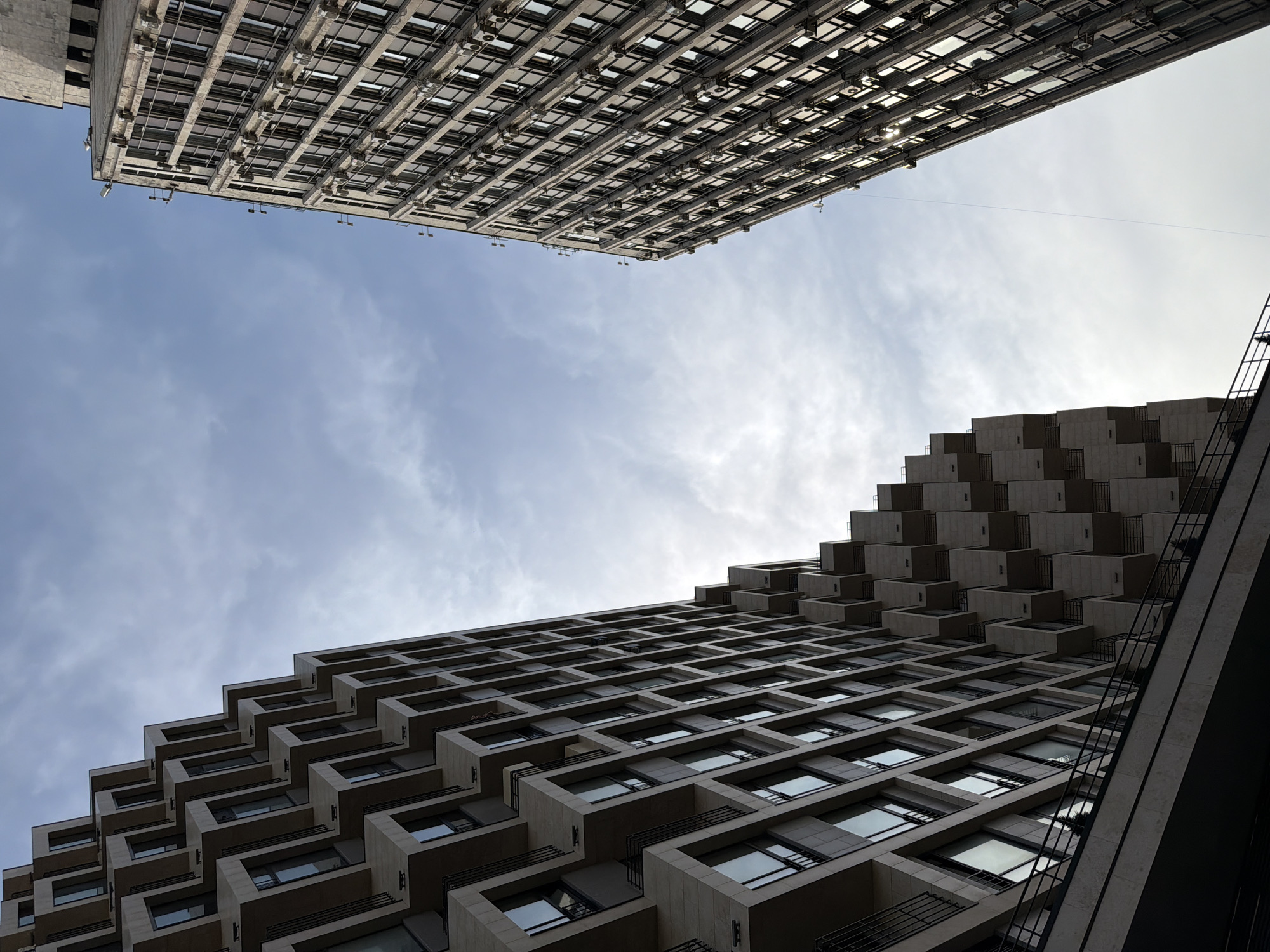 An upward, low-angle view of two high-rise buildings in Moscow, Russia, reveals a section of sky between them. The building at the top of the frame is a light-colored, rectangular structure featuring multiple rows of windows, each accompanied by a protruding air conditioning unit. The lower building is a light-colored structure with regularly recessed windows that are positioned in a stepped pattern as the building gets taller. A dark, vertical line is visible along the lower right side of the lower building. The sky is pale blue with thin wisps of clouds.