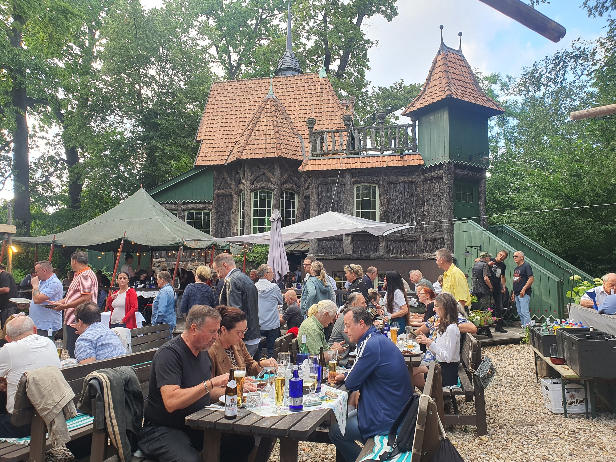The image depicts an outdoor gathering of people at a rustic, possibly historic, establishment in Falkensee, Germany, identified as the "Biergarten des alten Forsthauses." The architecture features a distinctive dark wood construction with a red-tiled, multi-peaked roof and green accents. In the foreground, several wooden picnic tables are occupied by diverse groups of individuals, engaged in eating, drinking, and conversation. Bottles of beer, glasses, plates of food, and cutlery are visible on the tables. The ground is covered in small, light-colored pebbles.
In the middle ground, a large, green, canvas tent is set up, providing shade over additional seating areas. Many people are seen milling about, standing, and socializing under the tent and around the tables. Some individuals are interacting with service staff or queuing at a counter. The background is dominated by lush green trees, indicating a wooded or park-like setting surrounding the venue. The lighting suggests it is daytime, possibly late afternoon, with dappled sunlight filtering through the leaves.
The attire of the people ranges from casual to slightly more formal, reflecting a relaxed atmosphere. The overall scene conveys a sense of community and enjoyment, typical of a beer garden environment. The presence of a wooden staircase leading up to a balcony area on the building, along with decorative wooden railings, adds to the rustic charm of the location.