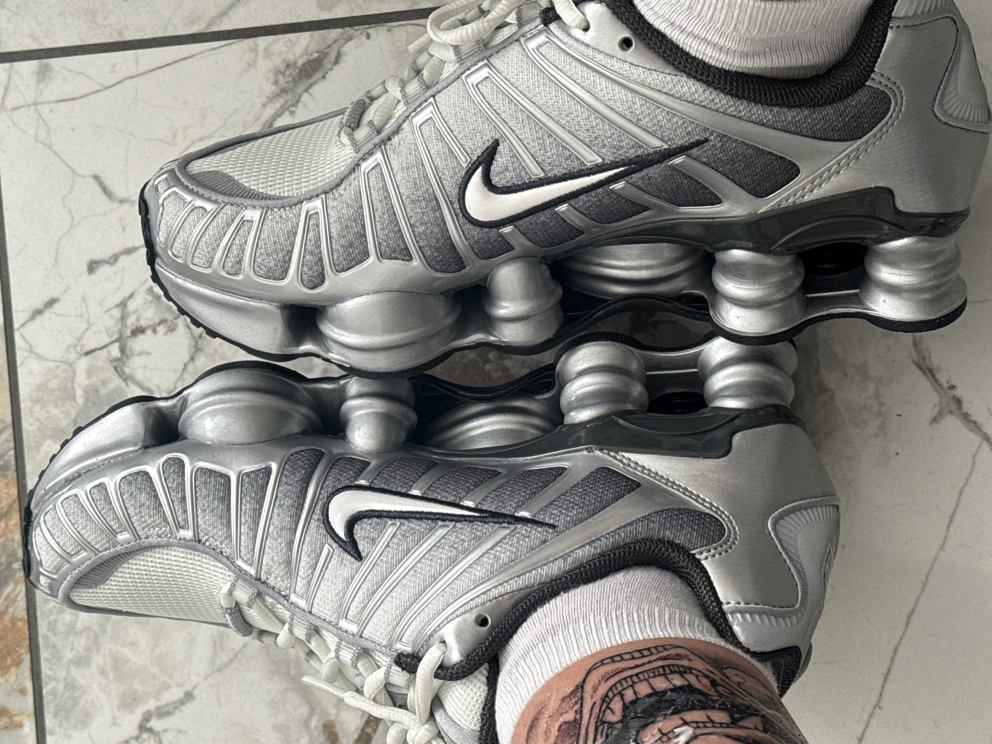 The image shows a pair of silver Nike Shox TL sneakers. The shoes are lying on a tiled surface that appears to be indoors. One of the shoes is being worn, as evidenced by the white sock and part of a tattoo visible on the wearer's ankle. There are no explicit indicators that this image was taken in Burgas, Bulgaria.
