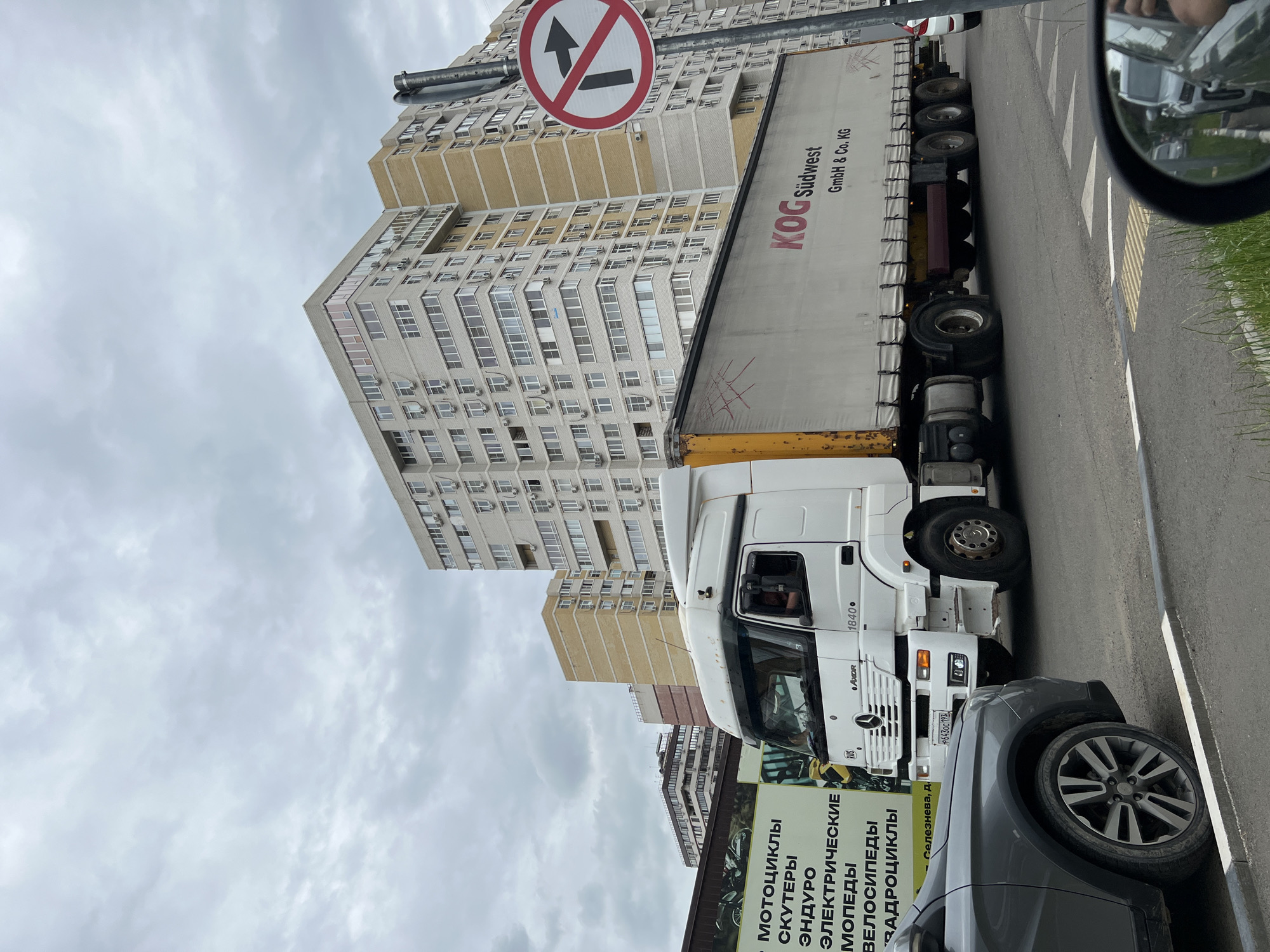 A white Mercedes-Benz Actros semi-truck with a white trailer is traveling on a road in Krasnodar, Russia. The truck is emblazoned with "KOG Südwest GmbH & Co. KG". To the left of the truck is a billboard advertising "МОТОЦИКЛЫ СКУТЕРЫ ЭНДУРО ЭЛЕКТРИЧЕСКИЕ МОПЕДЫ ВЕЛОСИПЕДЫ ЗАДРОЦИКЛЫ" (motorcycles, scooters, enduro, electric mopeds, bicycles, quad bikes) and a partial view of a gray car. In the background are tall, multi-story apartment buildings. A traffic sign prohibiting right turns is visible on the right side of the image. The sky is overcast with clouds.