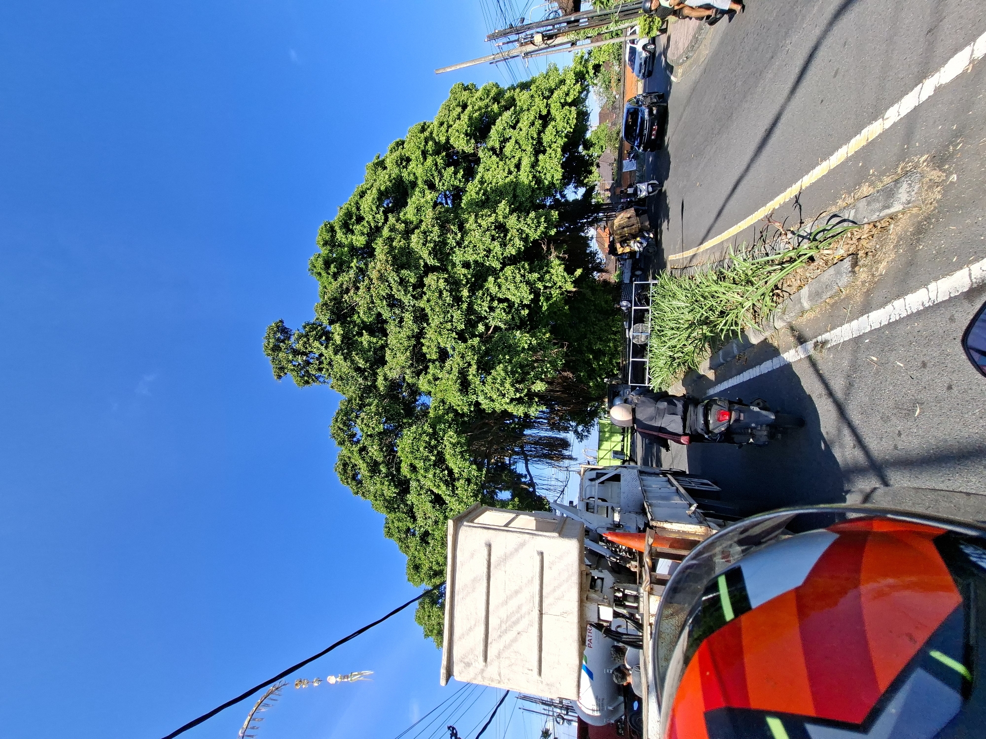 An eye-level, wide-angle outdoor shot captures a street scene in Banjarangkan, Indonesia, on a bright, clear day under a deep blue sky. Dominating the background is a massive, mature tree with a sprawling canopy of vibrant green leaves, its aerial roots dangling down. In the foreground, the perspective is from a motorcycle, with a red, black, and white patterned helmet visible. To the left, a white truck with a large, light-colored rectangular container on its back is parked or moving slowly. A white tanker truck with "PATRA" visible on its side is behind it. A person in a black jacket and helmet is riding a black scooter in the middle of the road, moving away from the camera. Further down the road, a black SUV and a white car are visible, along with other vehicles and pedestrians on the right side of the street. The road has a distinct yellow dividing line and white lane markings. Lush green plants line the median strip. Electrical wires and poles are present on the right side of the image, and a traditional Balinese decorative element hangs from a wire on the left.