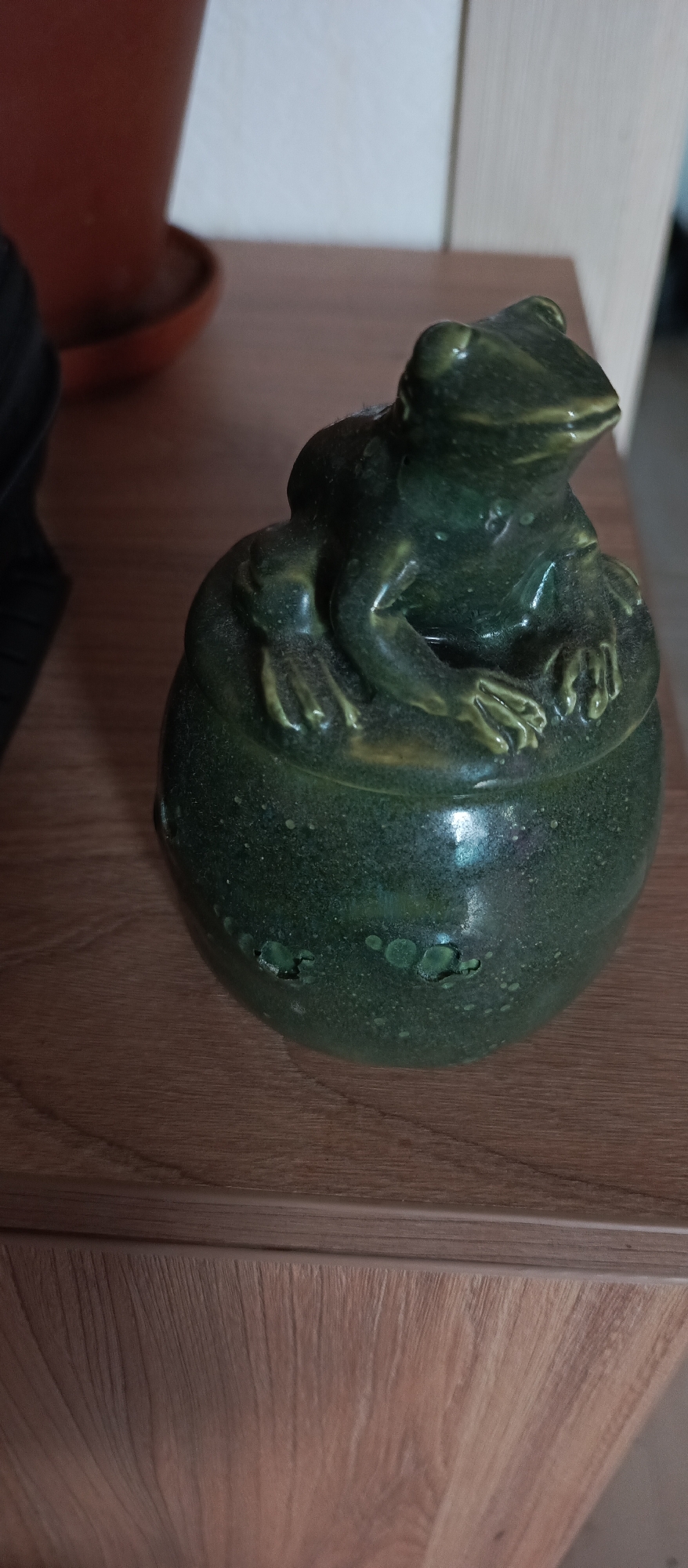 A dark green, glazed ceramic frog sits atop a matching pot, which rests on a wooden surface. The frog is depicted in a seated position with its front legs resting on the rim of the pot. The pot has a slightly irregular, bumpy texture with some lighter green circular embellishments. The wooden surface appears to be a table or shelf with a visible wood grain. In the background, a portion of a reddish-brown pot and a white wall are visible. The image is taken from a slightly high angle, focusing on the ceramic frog and pot. There are no people or animals present in the image. This scene is not specific to Bugry, Russia.