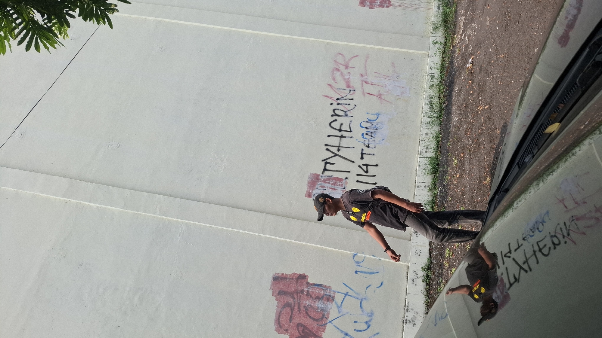 A young man wearing a dark t-shirt, dark baseball cap, and grey jeans walks along a white wall covered in graffiti. The graffiti includes red spray paint markings and black lettering that appears to say "TYHERINZR" and "114 TEARU". A reflection of the man and some of the graffiti is visible on the hood of a car in the foreground, which is parked on a dirt and gravel surface. Green foliage is visible at the top right of the image, suggesting an outdoor setting with vegetation. The wall appears to be a structural element, possibly part of a building or a boundary. The overall scene suggests an urban environment, possibly in a less maintained area, with evidence of youth culture through the graffiti. The man's posture indicates he is walking, with his right hand slightly raised. There are no other people or distinct animals visible in the frame.