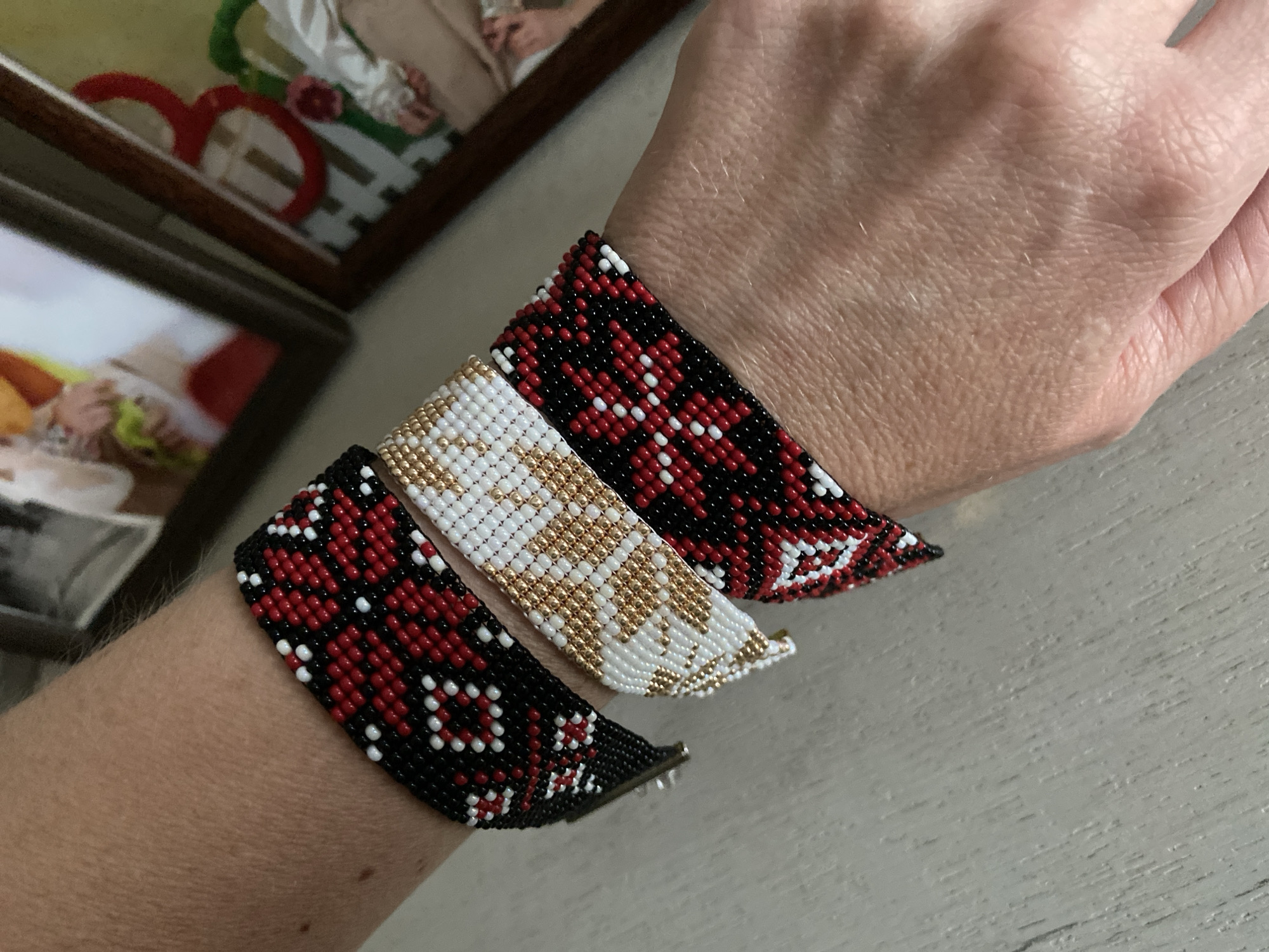 The image displays a close-up view of a person's forearm and wrist, featuring three handcrafted beaded bracelets. The bracelets are made from small seed beads, each exhibiting a unique geometric or floral pattern. The leftmost bracelet is predominantly black with red and white patterned beads, and a metal clasp is visible. The central bracelet features white and golden beads in a distinct pattern, while the rightmost bracelet is primarily red with black and white patterned beads. The patterns on all bracelets appear to be intricate and stylized. The arm rests on a light-colored, textured surface, possibly painted wood. In the blurred background, two framed items are visible; one contains an indistinct image with prominent red heart-shaped glasses, and the other shows a further out-of-focus image. The scene suggests an indoor setting with soft, ambient lighting. No specific activity beyond showcasing the bracelets is evident, and no text is visible.