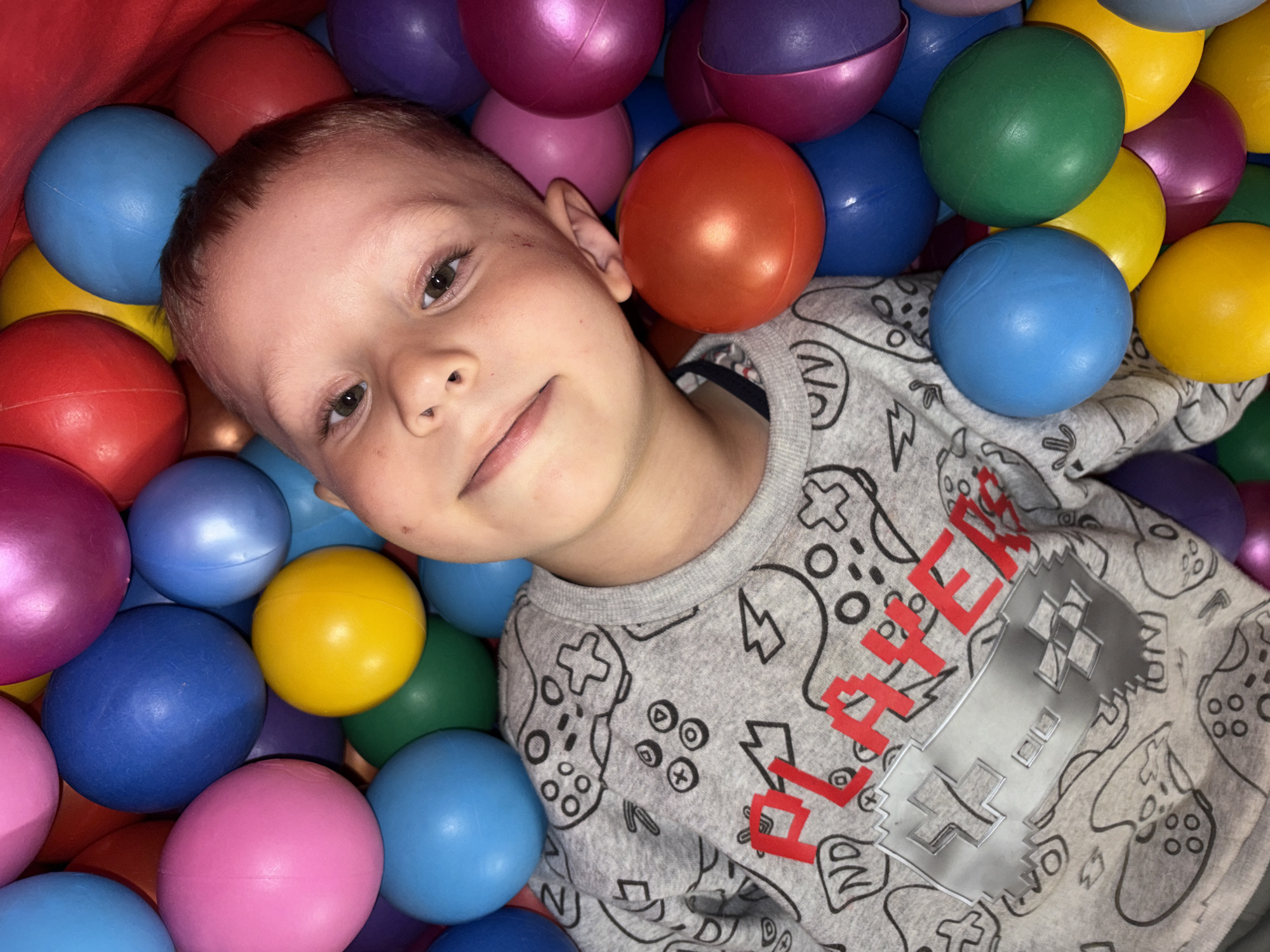 A young boy is depicted in a close-up, lying in a vibrant ball pit. He is positioned slightly diagonally in the frame, looking directly at the camera with a calm, content expression and a subtle, soft smile. His eyes are open and engaging.
The boy has short, light brown hair. He is wearing a grey long-sleeved shirt patterned with outlines of various video game controllers in black. On the front of his shirt, the word "PLAYER" is visible in a pixelated red font, alongside a large, metallic silver, pixelated game controller graphic.
He is surrounded by a dense collection of colorful plastic balls, which fill the entire image. The balls come in a variety of hues, including red, blue, pink, purple, yellow, orange, and green, creating a bright and playful environment. The lighting appears to be artificial and warm, highlighting the smooth, reflective surfaces of the balls and the boy's skin. The setting suggests an indoor play area, likely a home or entertainment center, located in Chaikovsky, Russia. The image captures a moment of relaxed and happy playtime.