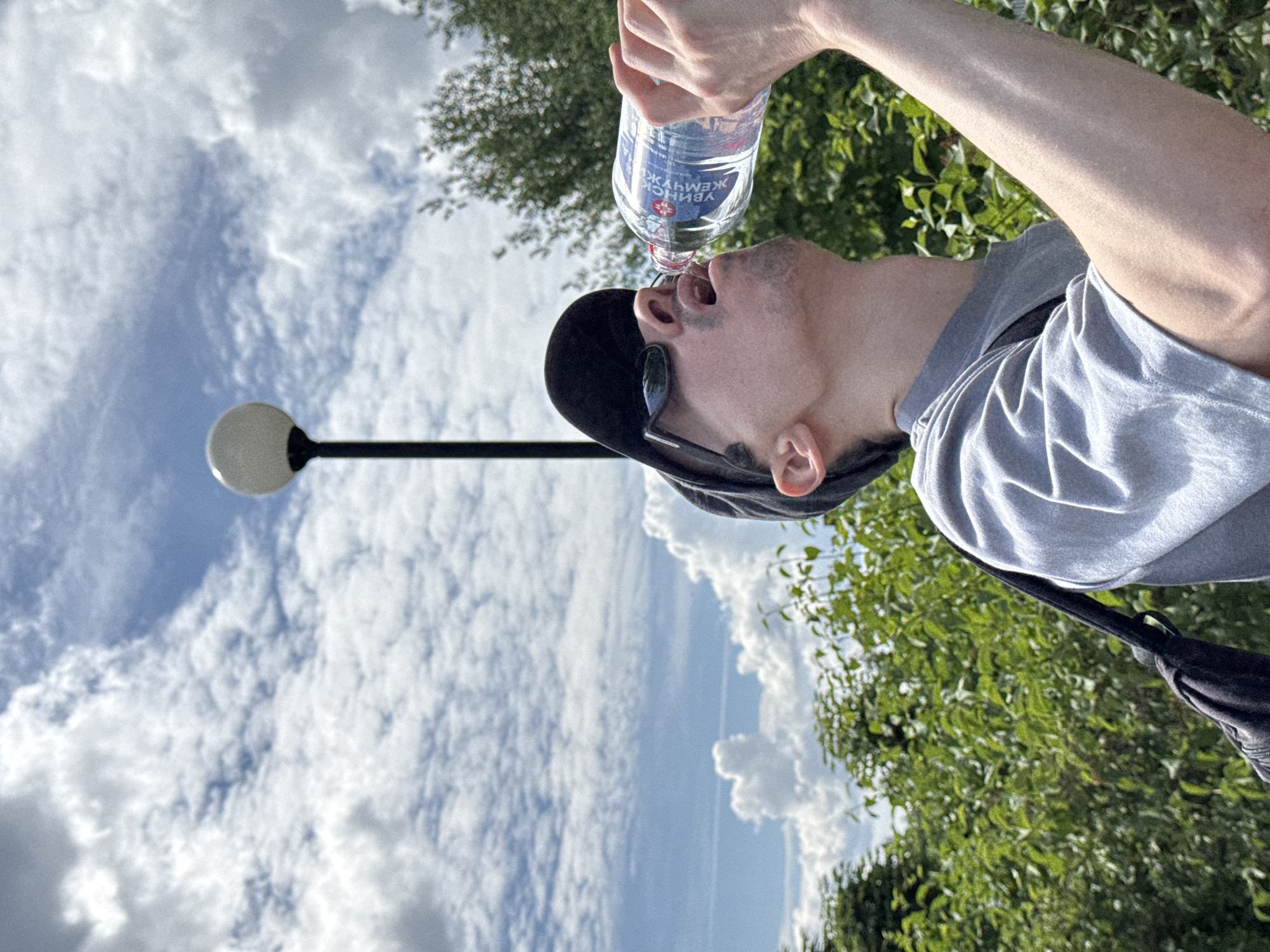 A lone male individual is depicted outdoors, seen from the mid-chest up, drinking from a plastic water bottle. He wears a grey short-sleeved t-shirt, a black baseball cap, and dark sunglasses. His head is tilted back, with his mouth open to the bottle, held by his raised right hand. A black backpack strap is visible on his left shoulder. The water bottle label is blue and white, prominently displaying "УВИНСКИЙ ЖЕМЧУГ" (Uvinsky Zhemchug), a brand of water produced in the Udmurtia Republic, Russia. In the background, a tall black street light with a spherical white fixture is positioned to the left. The sky is bright blue with scattered white cumulus clouds. Lush green foliage, consistent with bushes and trees, forms the backdrop. The scene suggests a sunny day in an urban park or public green space, specifically located within the city of Izhevsk, Russia, inferred from the locally sourced water bottle brand.