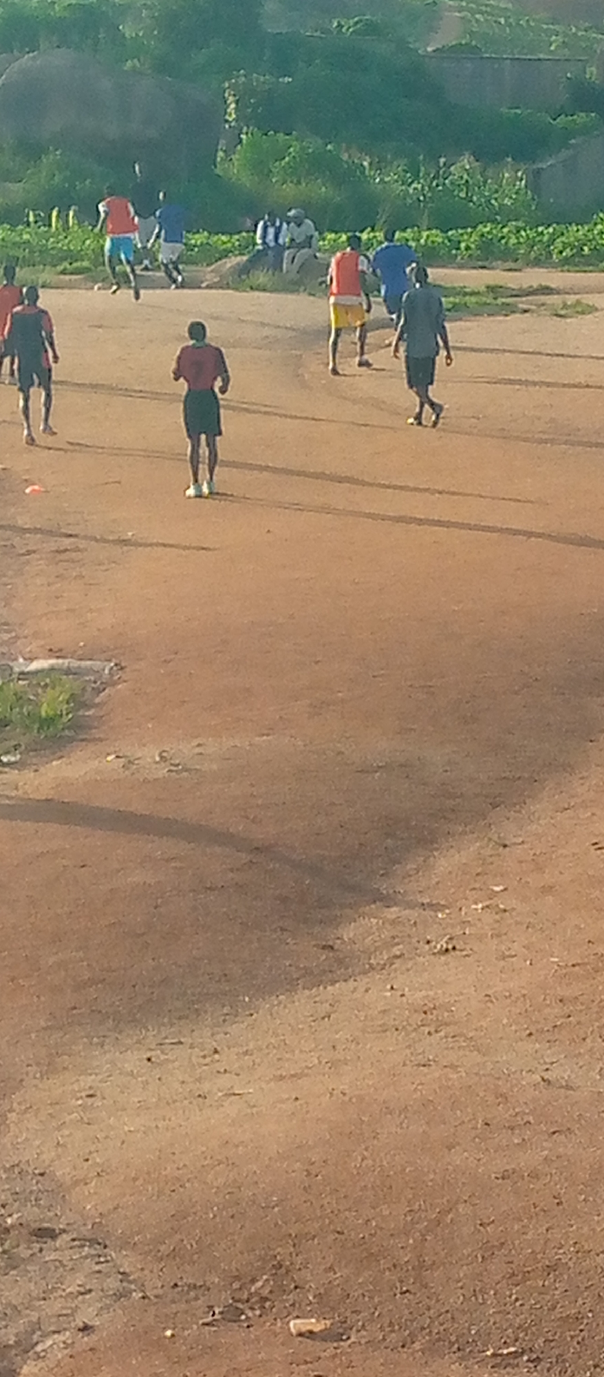 The image depicts an outdoor scene of a group of young men playing soccer on a dirt field. The field is predominantly brown, with sparse patches of green grass and vegetation visible in the background. A rocky, overgrown hill rises in the distance, with some structures or walls made of stone or concrete. Several individuals are captured in motion, suggesting active gameplay. Some are wearing brightly colored athletic jerseys, primarily red and orange, while others are in darker attire. The lighting indicates it is likely late afternoon or early morning, casting long shadows on the ground. The overall atmosphere suggests a casual sporting event in a somewhat undeveloped, rural or semi-rural setting.