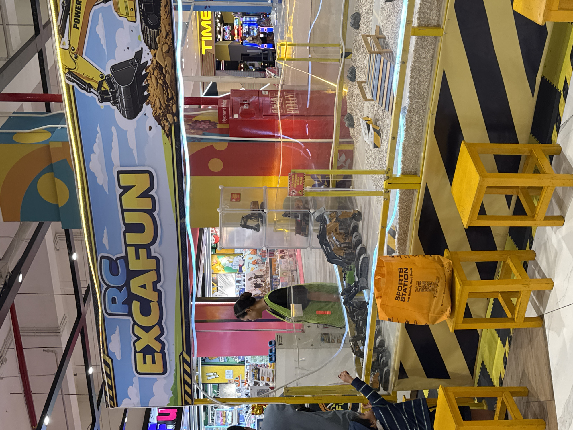 The image shows an indoor excavator game called "RC EXCAFUN" in a retail environment, likely a shopping mall in Kuta, Indonesia. The game area features a small excavator model operating within a fenced enclosure filled with gravel. The walls of the enclosure are partially transparent. Adjacent to the play area are black and yellow warning-striped panels, and several yellow wooden chairs.
A woman wearing a green vest and dark hair is present within the excavator game's area. Two individuals, including a child wearing a striped shirt, appear to be engaged with the game or watching. The background reveals additional retail spaces and signage, including a "TIME" sign. A "SPORTS STATION" branded bag is placed on one of the chairs. Signage in the background might have Indonesian text.