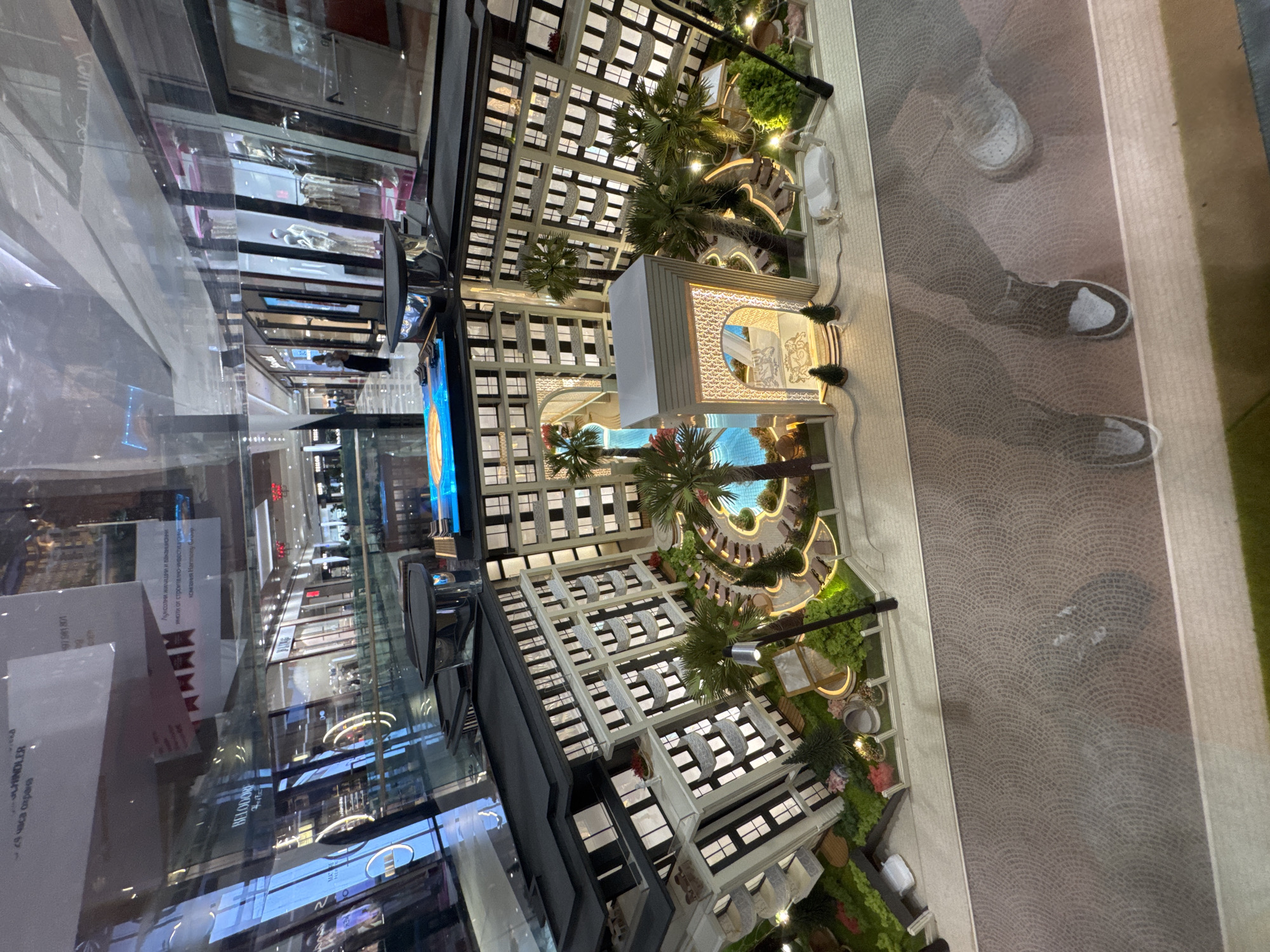 The image shows a model of a luxury residential complex, likely displayed in a shopping mall. The model features a tall building complex with a pool area, a decorative pavilion, landscaping, and street paving. The reflection in the glass displays of the mall suggests an upscale shopping center location, possibly in Sofia, Bulgaria, as indicated by the Cyrillic text on nearby advertisements. People are visible as reflections in the background.