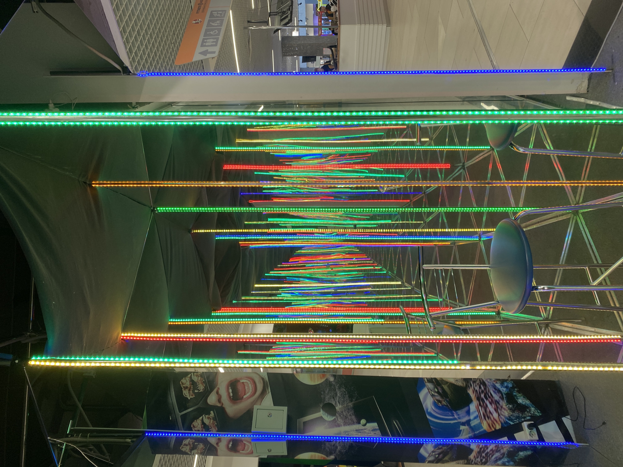 The image depicts a long tunnel of mirrors, lined with colorful LED strips of blue, green, red, and yellow light, creating an infinity effect. In the foreground, there is a bar stool with a blue seat and metal legs. At the tunnel's entrance, there is an advertisement with a person's mouth open. The photo was taken inside the shopping mall "Talismann" in Izhevsk, Russia.