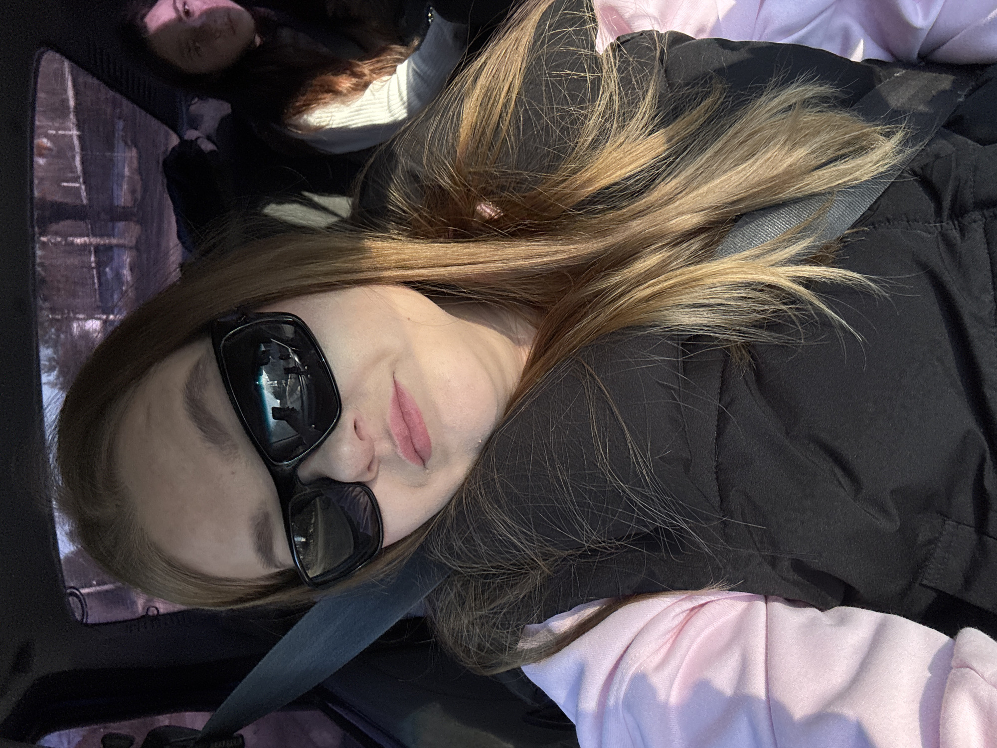 A fair-skinned individual with long, highlighted blonde hair is seen wearing black sunglasses and a black padded jacket, with a seatbelt visible across their body. They appear to be in a vehicle. A person with long brown hair is partially visible in the upper left corner of the image, likely in the back seat. Outside the car window, blurred buildings or trees are discernible. Due to the lack of specific identifying landmarks, pinpointing the exact location within Chaikovsky, Russia, is not possible. The image depicts a person wearing sunglasses and a seatbelt in a car with another person.
