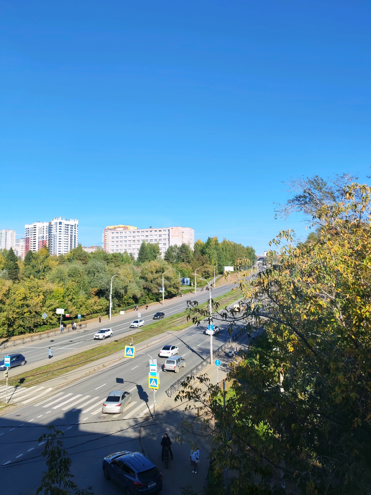 This is a daytime, high-angle, eye-level shot of a city street in Izhevsk, Russia, on a clear day with a bright blue sky. The scene depicts a multi-lane road with several cars in motion, including a silver sedan, a white SUV, and a black sedan. Pedestrians are visible on the sidewalks and crossing the street at a marked crosswalk. Yellow pedestrian crossing signs are present. On the right side of the road, there are tram tracks. Beyond the road, there is a dense line of trees with autumn foliage, displaying a mix of green, yellow, and brown leaves. In the background, large multi-story residential buildings are visible, one white and blue, and another a pale pinkish-yellow. The overall impression is of an urban environment with elements of nature, likely during the autumn season.