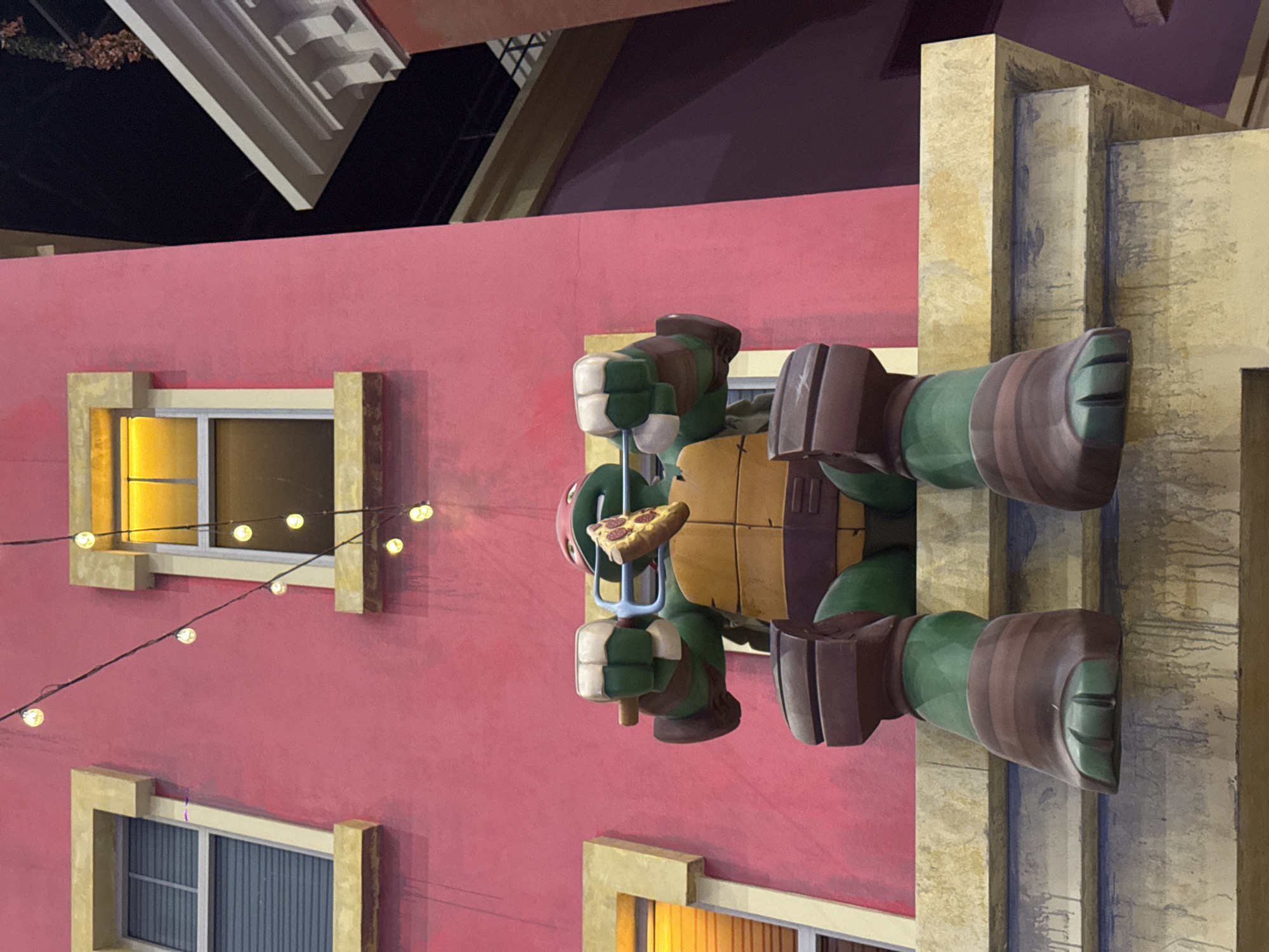 The image appears to be a street scene with a Teenage Mutant Ninja Turtle figure holding a slice of pizza. The background features a pink building with windows and string lights. The architectural style suggests a Mediterranean or European influence. Based on the presence of the Teenage Mutant Ninja Turtle figure and the stylized building, it is likely a themed area or attraction, possibly a children's play area. It is not possible to confirm the location in Moscow, Russia, without more information.