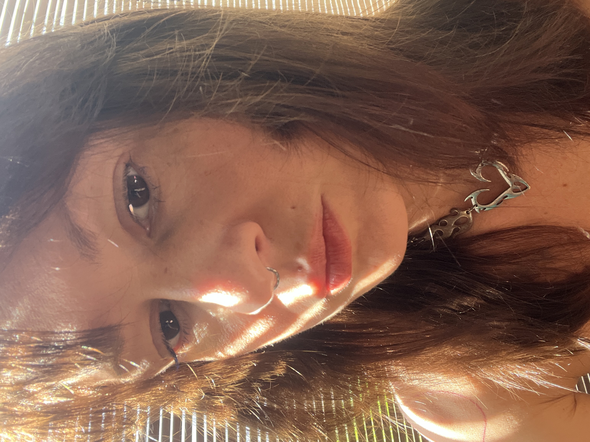A close-up, slightly rotated, portrait shot of a woman with medium-length brown hair. Her skin appears fair, and she has a nose ring on her left nostril. She is wearing an earring shaped like a heart with additional decorative elements. Sunlight is prominently featured, creating strong highlights and shadows across her face. The background is out of focus but appears to include vertical lines, possibly from blinds or a fence. The location in North Kuta, Indonesia is not discernible from the visual information provided. No actions or interactions are visible.